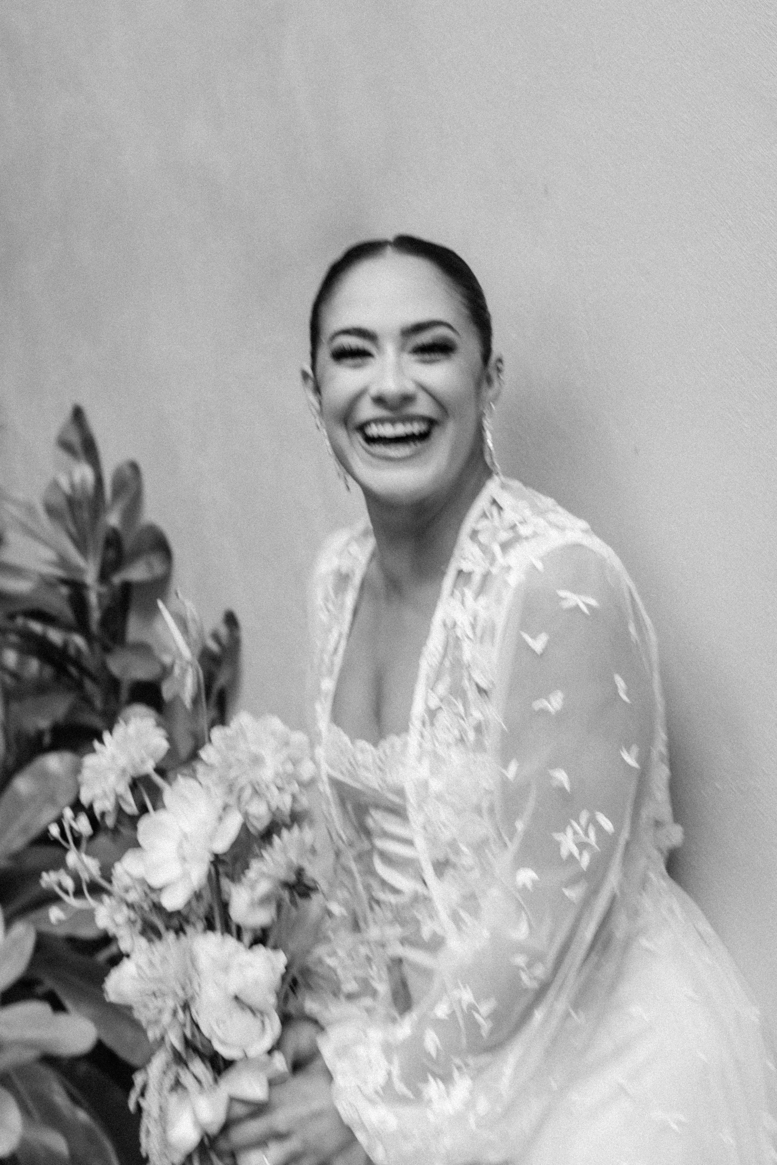 Black and white photo of a smiling woman in a dress, holding a bouquet of flowers, standing in front of a plain wall.