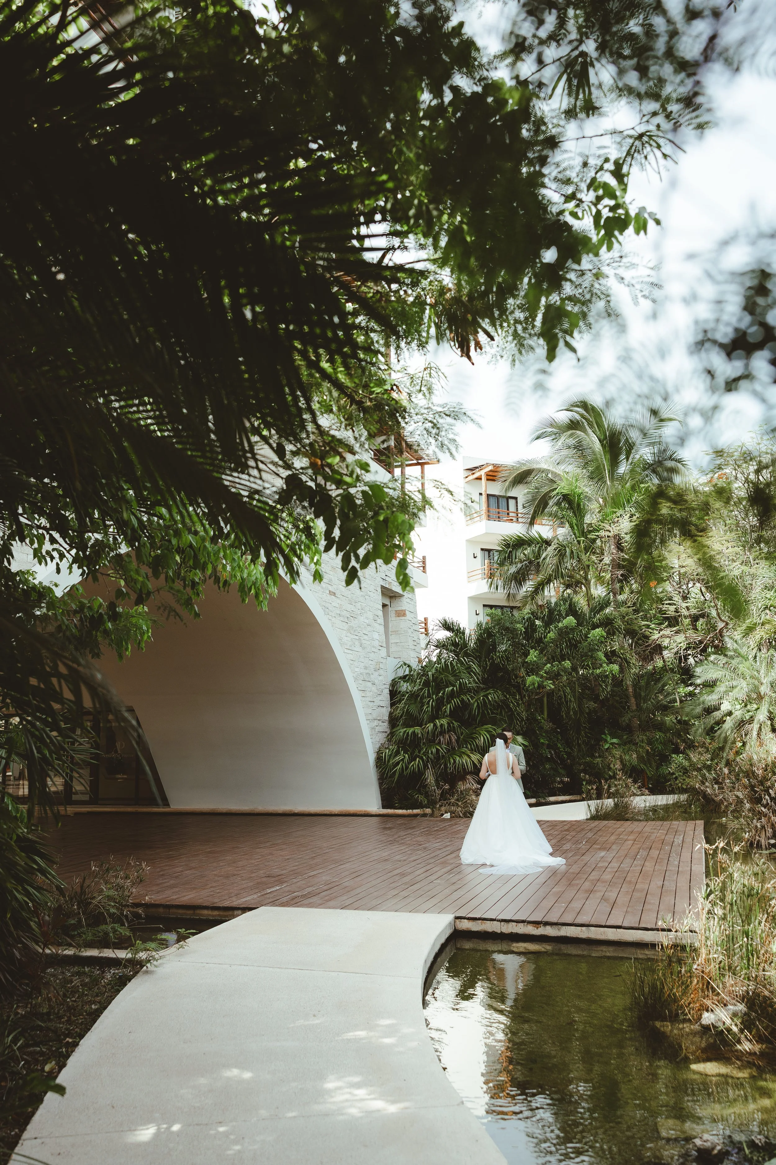 A bride in a white wedding dress walking on a wooden deck surrounded by lush green trees and plants, near a pond with a curved concrete walkway.