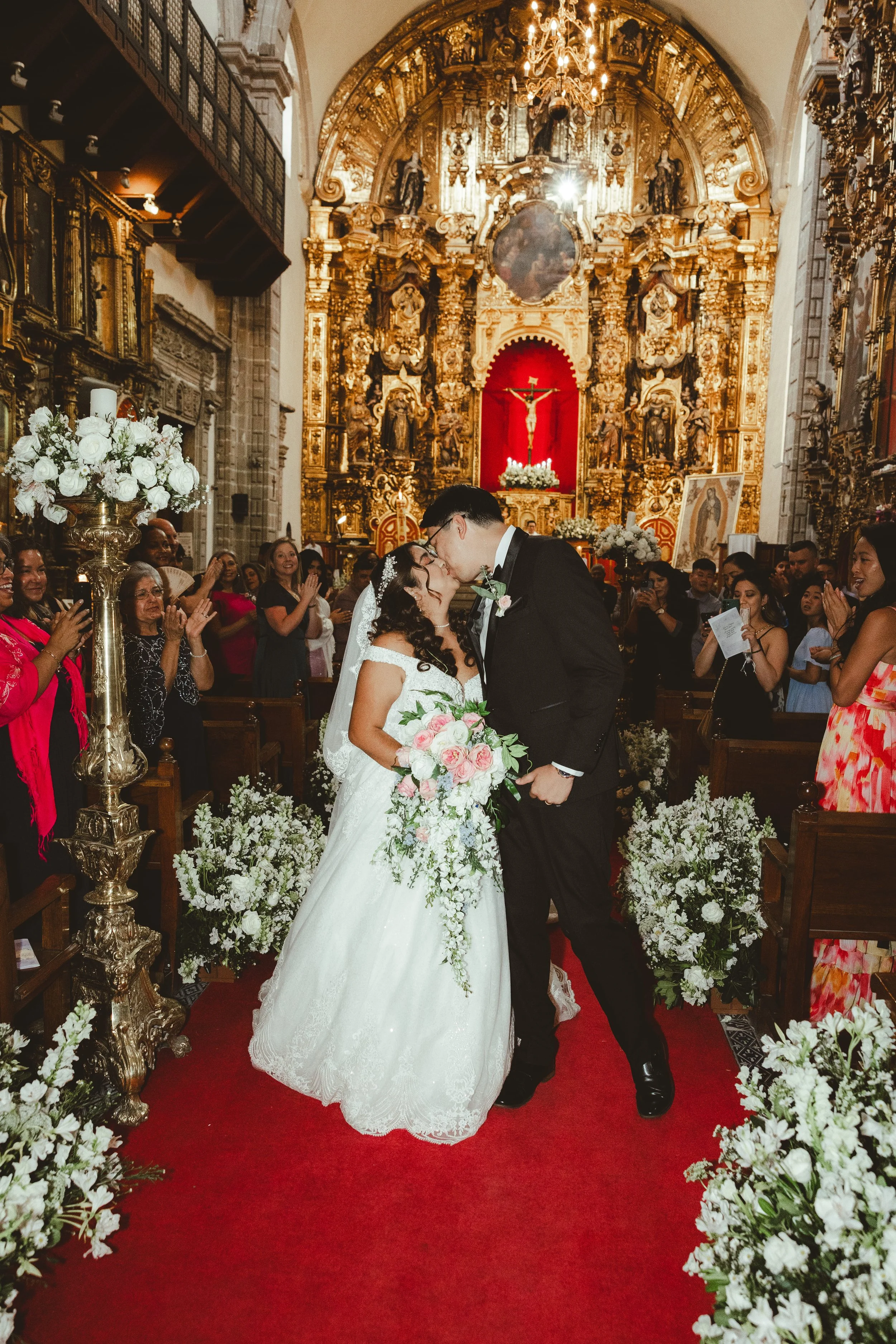 A newlywed couple sharing a kiss at the altar in a church decorated with white flowers, gold accents, and a red backdrop behind a crucifix, with guests smiling and clapping in the background.