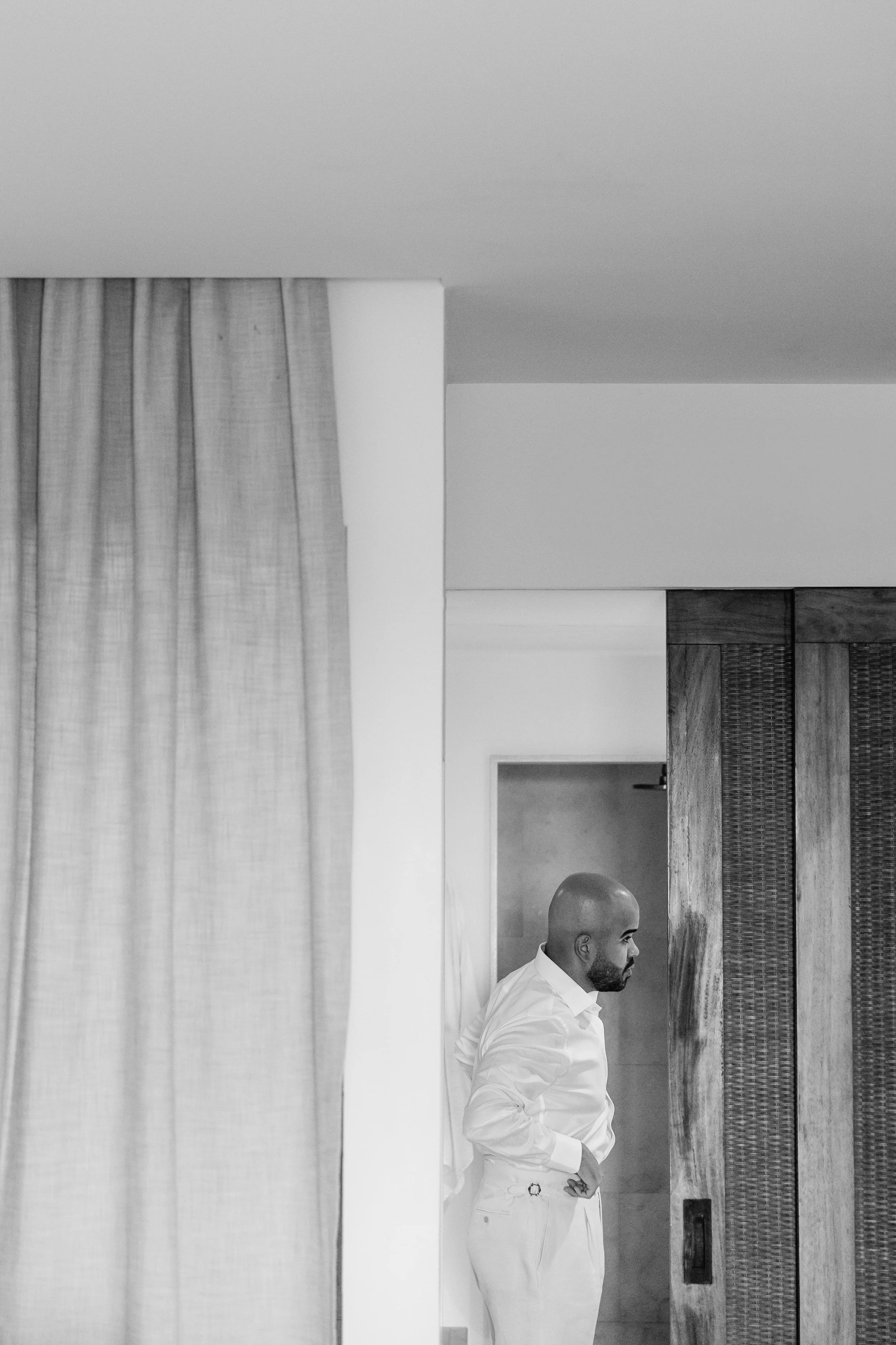 Man with a shaved head, beard, in white dress shirt and pants, standing indoors near a wooden partition and a curtain.