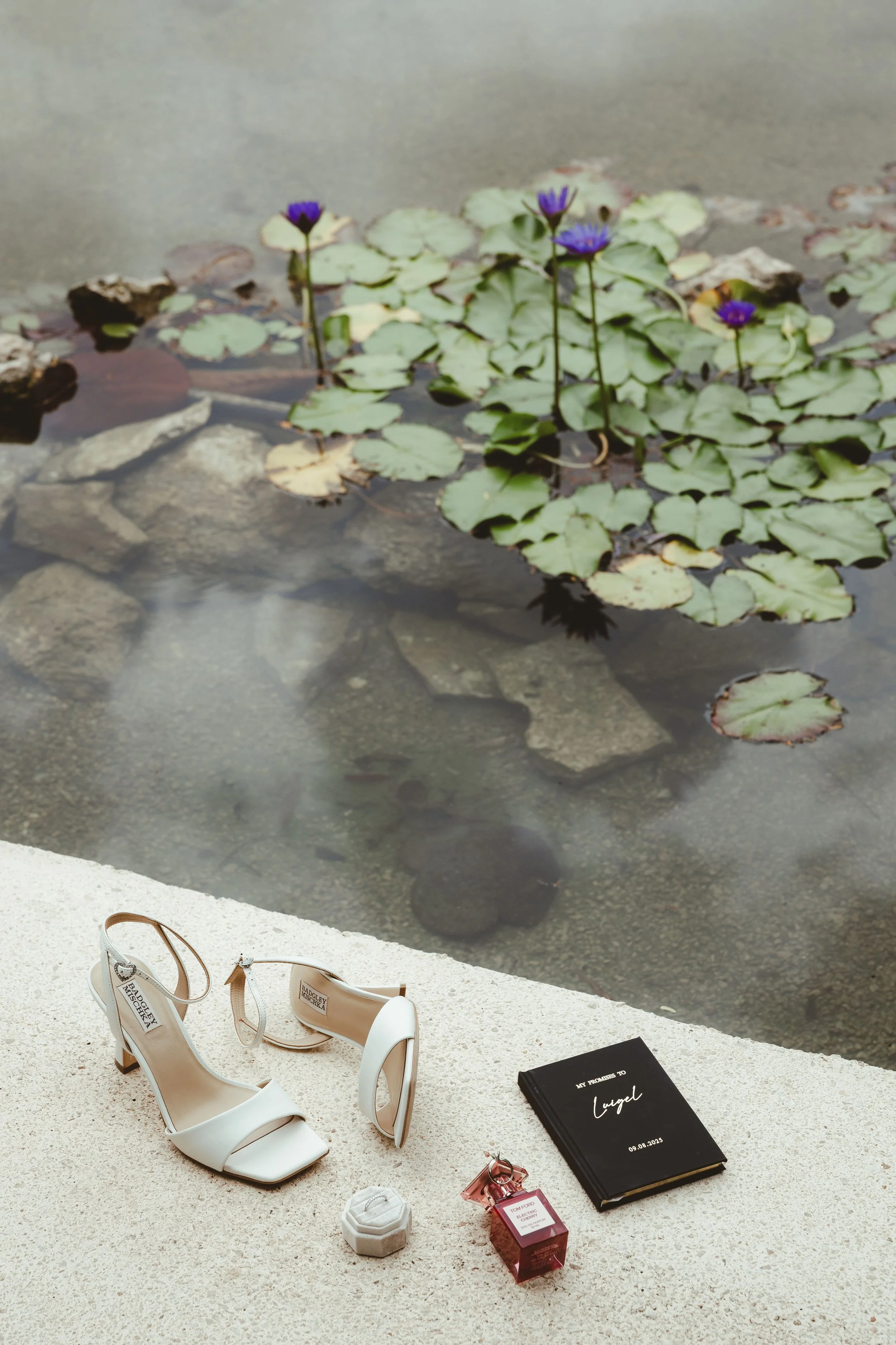 A white high-heeled sandal shoe on a white surface with fashion items surrounding it, including a black notebook titled "My Promis to Angel," perfume bottles, and jewelry. The background shows a pond with lily pads and purple water lilies.