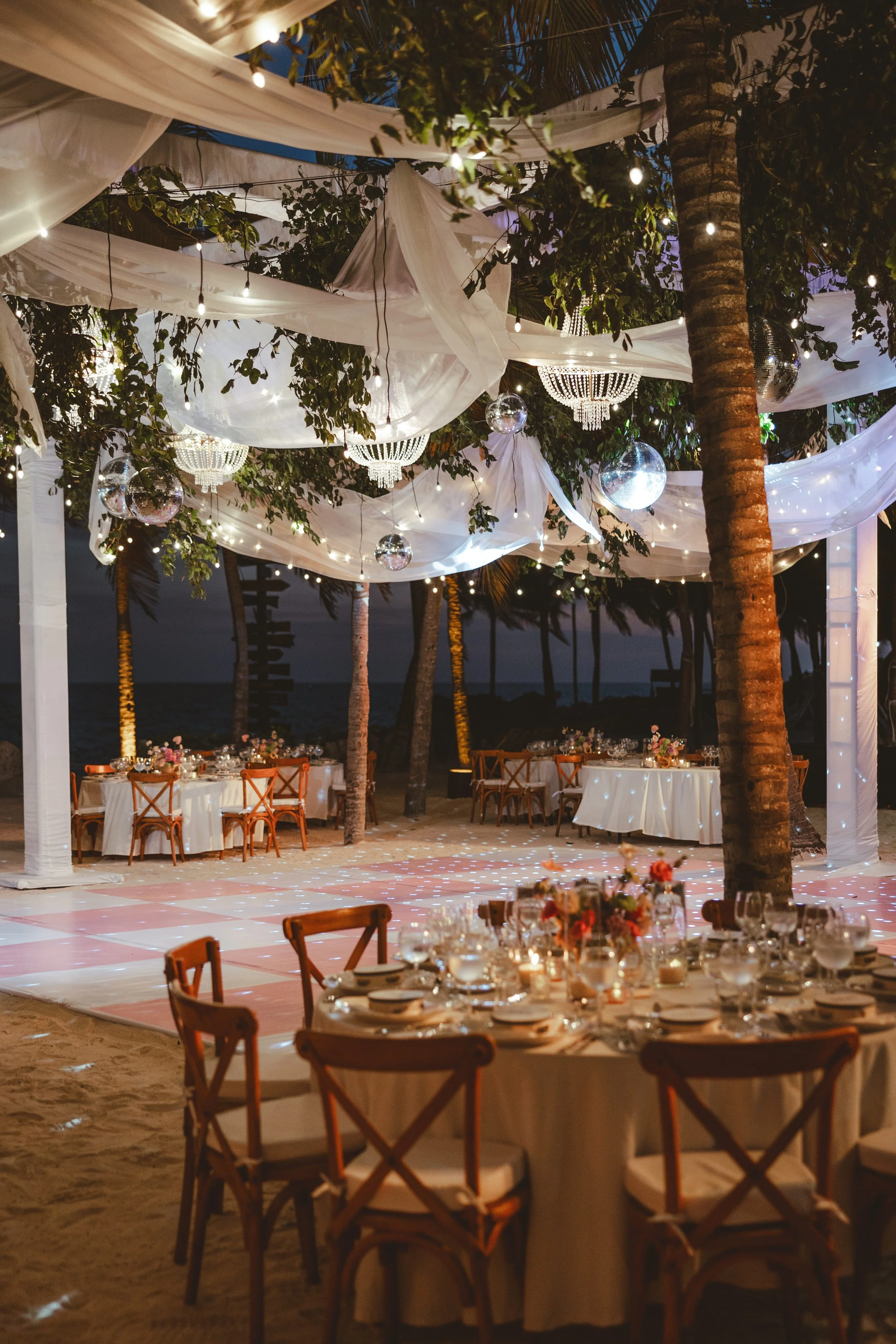 Outdoor event setup at night with decorated tables, string lights, hanging disco balls, palm trees, and flowing fabric drapes in a beach setting in Xcaret Wedding.