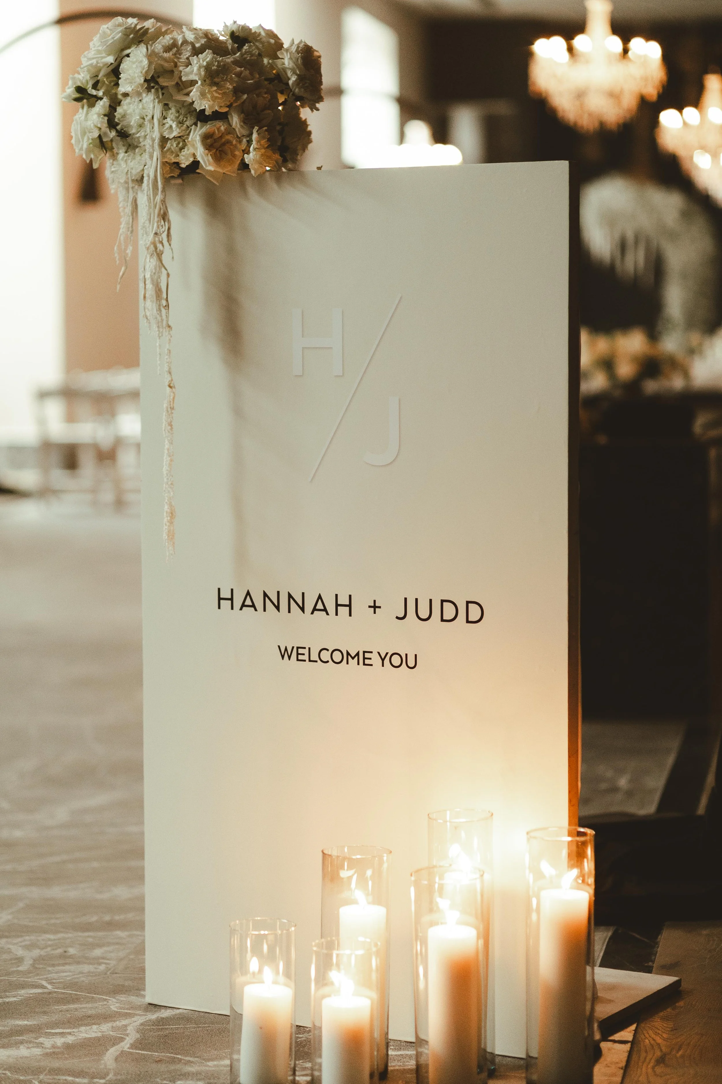 A wedding welcome sign with the words 'Hannah + Judd Welcome You,' decorated with a floral arrangement at the top left corner and surrounded by lit candles in glass holders. The background shows a chandelier and a softly lit interior.