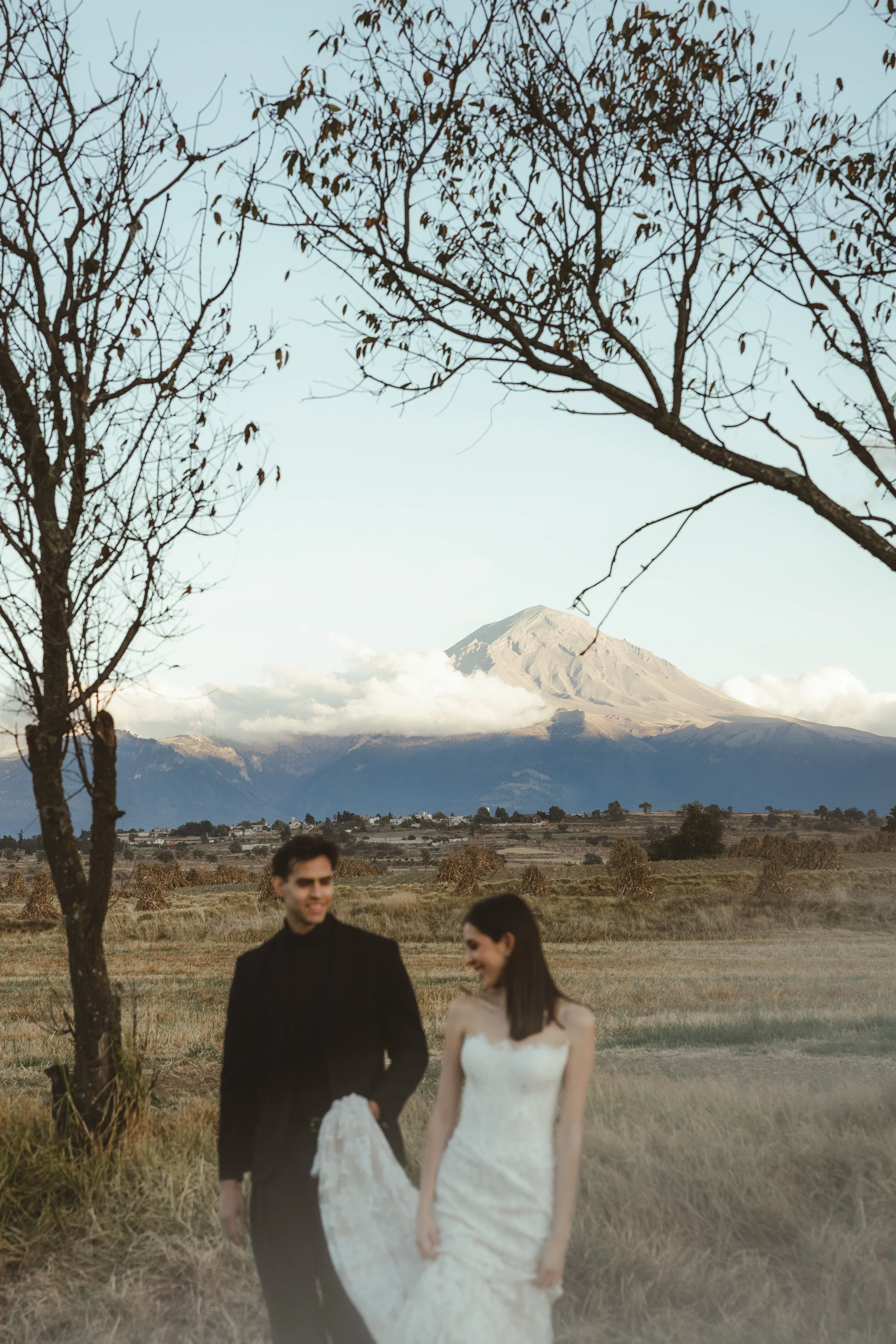 A couple dressed in formal wedding attire holding hands outdoors with a mountain and partly cloudy sky in the background.