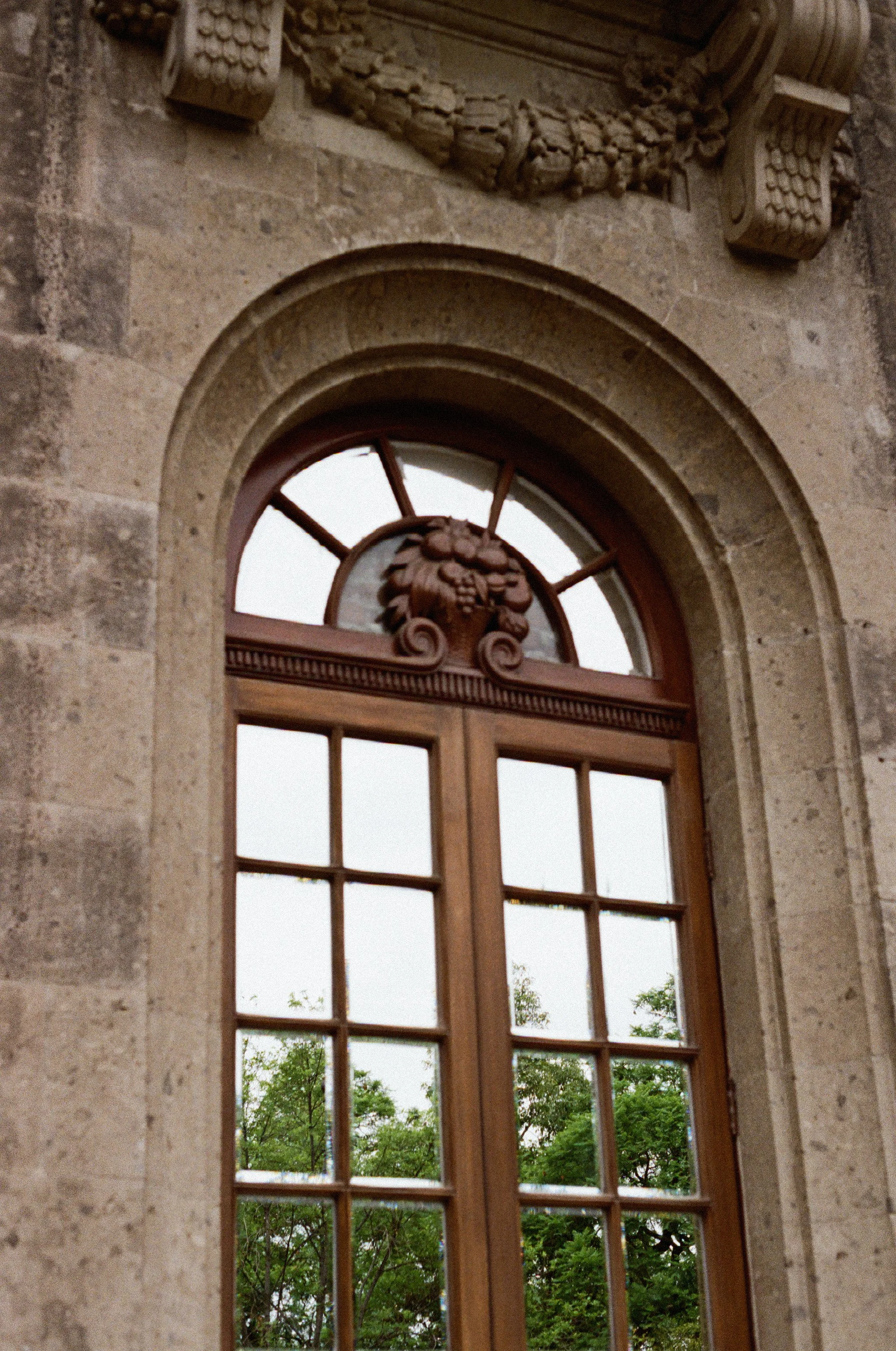 A window with an arched top, decorative woodwork, and a view of green trees outside, set in a stone wall with intricate stone carvings.