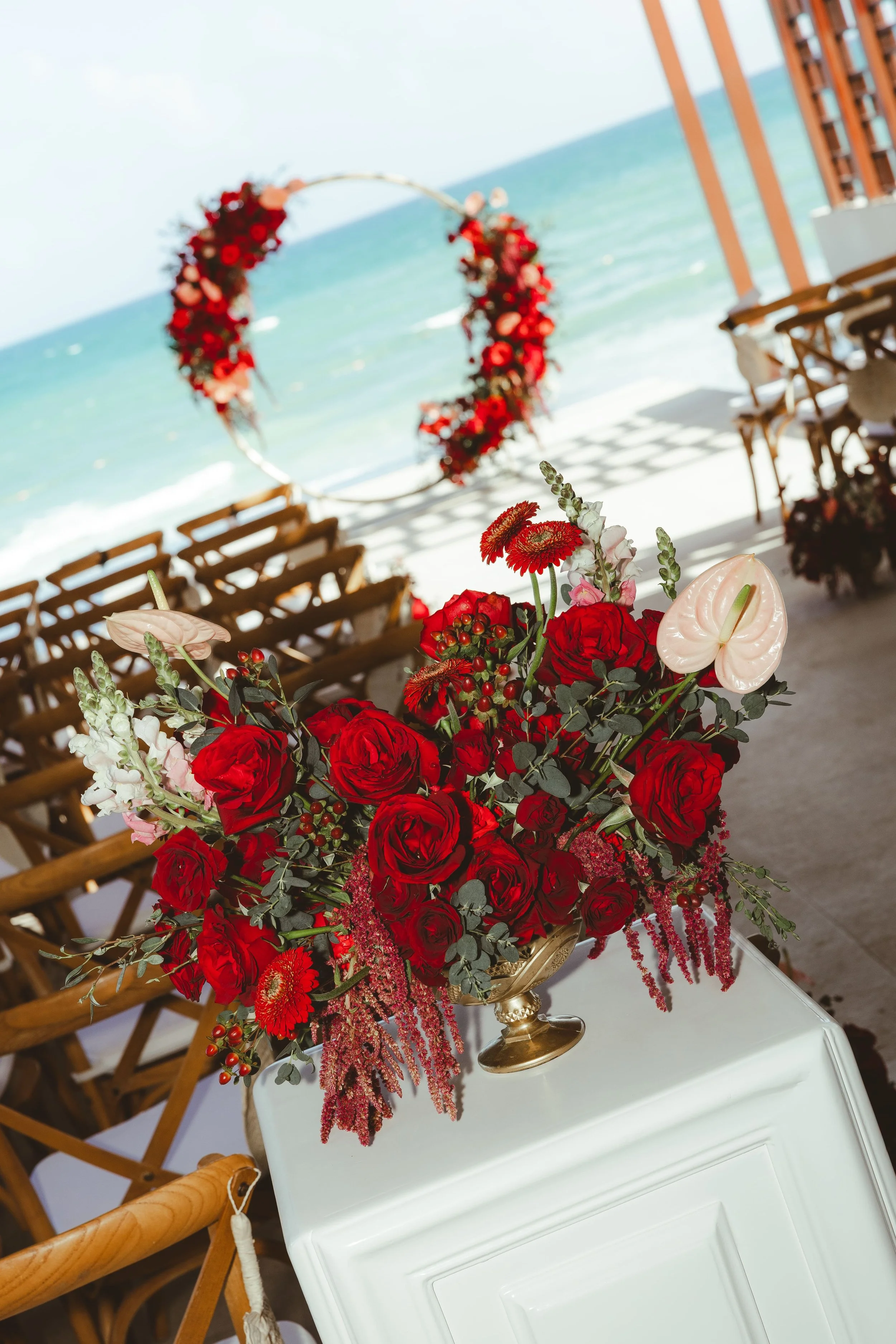 A large floral arrangement featuring red roses, white anthuriums, and other red and white flowers in a gold vase, placed on a white table with chairs and an outdoor beach scene in the background.
