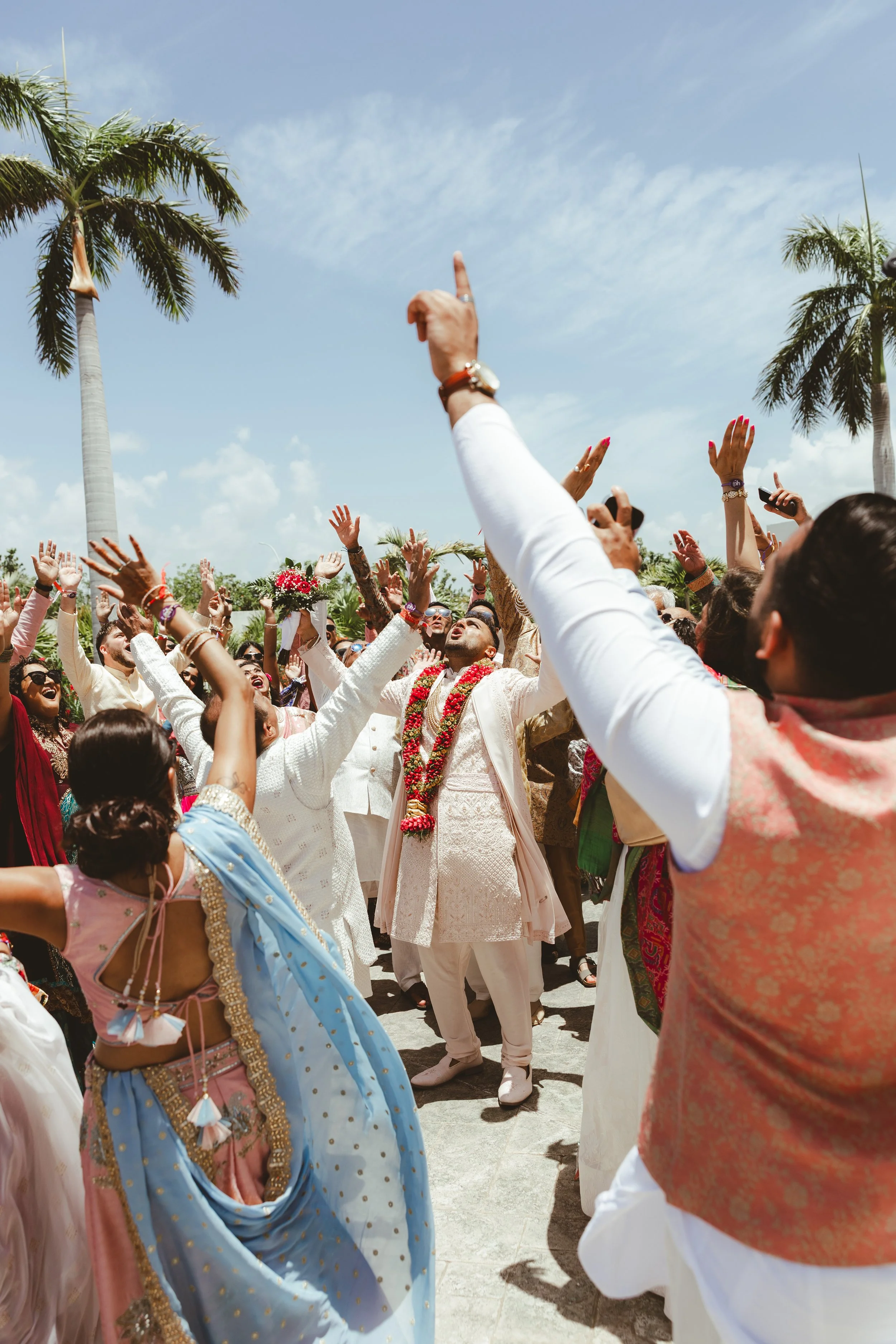 A joyful outdoor wedding celebration with people dressed in traditional Indian attire, raising hands in the air, with palm trees and a blue sky in the background.