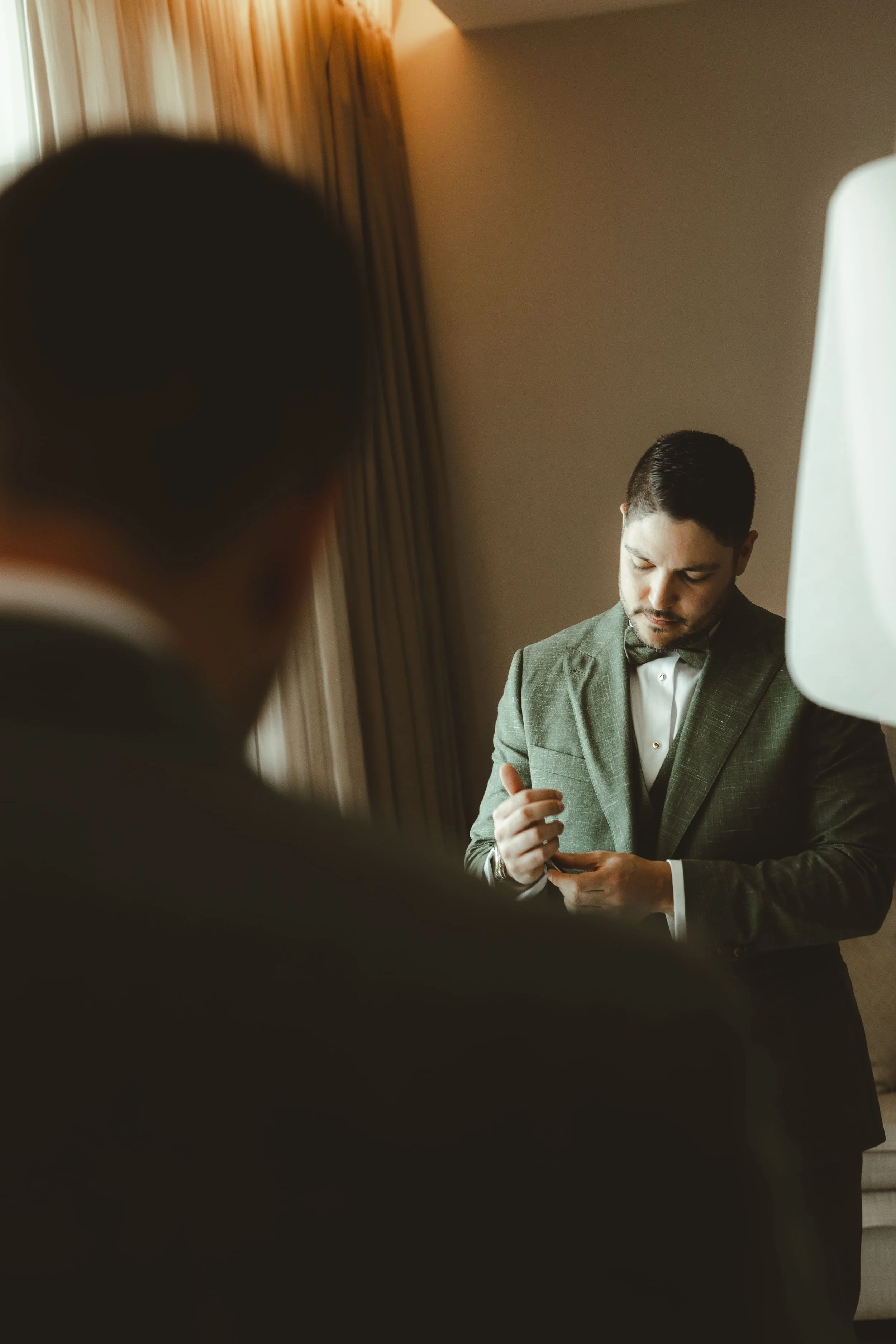 A man dressed in a suit looking down, with another person or object in the foreground, partially obstructing the view. The background features a beige wall and curtains.