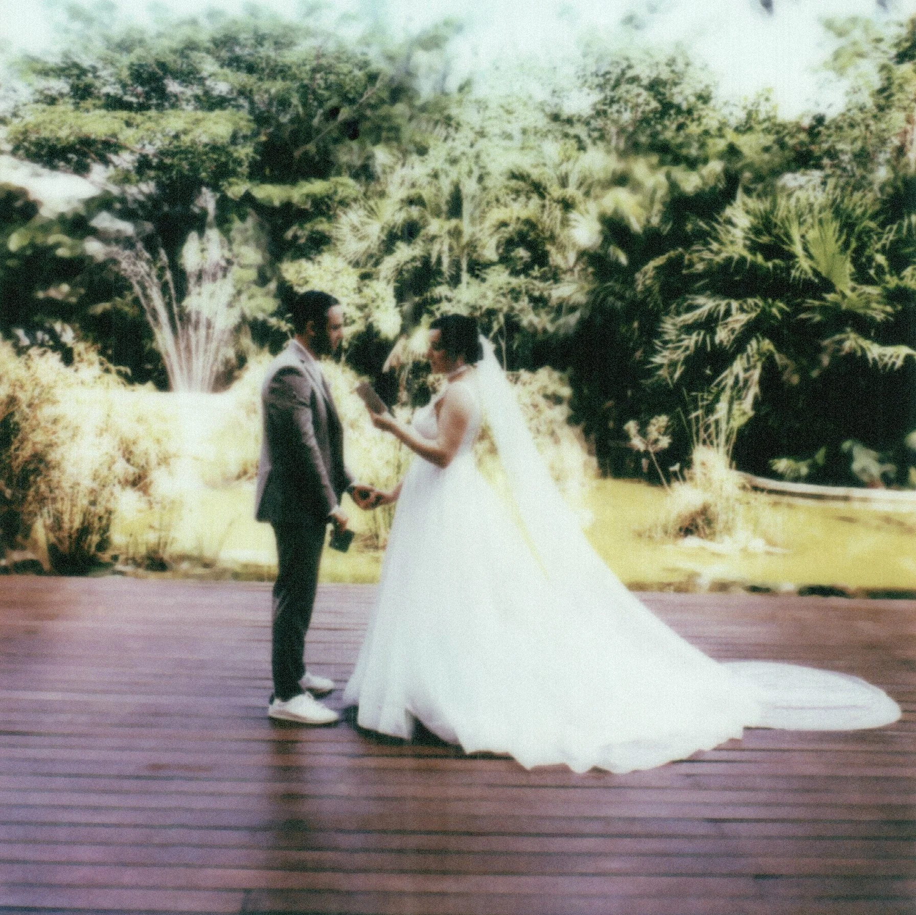 A couple holding hands during their wedding ceremony outdoors in a garden with lush green trees and bushes, with the bride wearing a white dress and the groom in a suit.