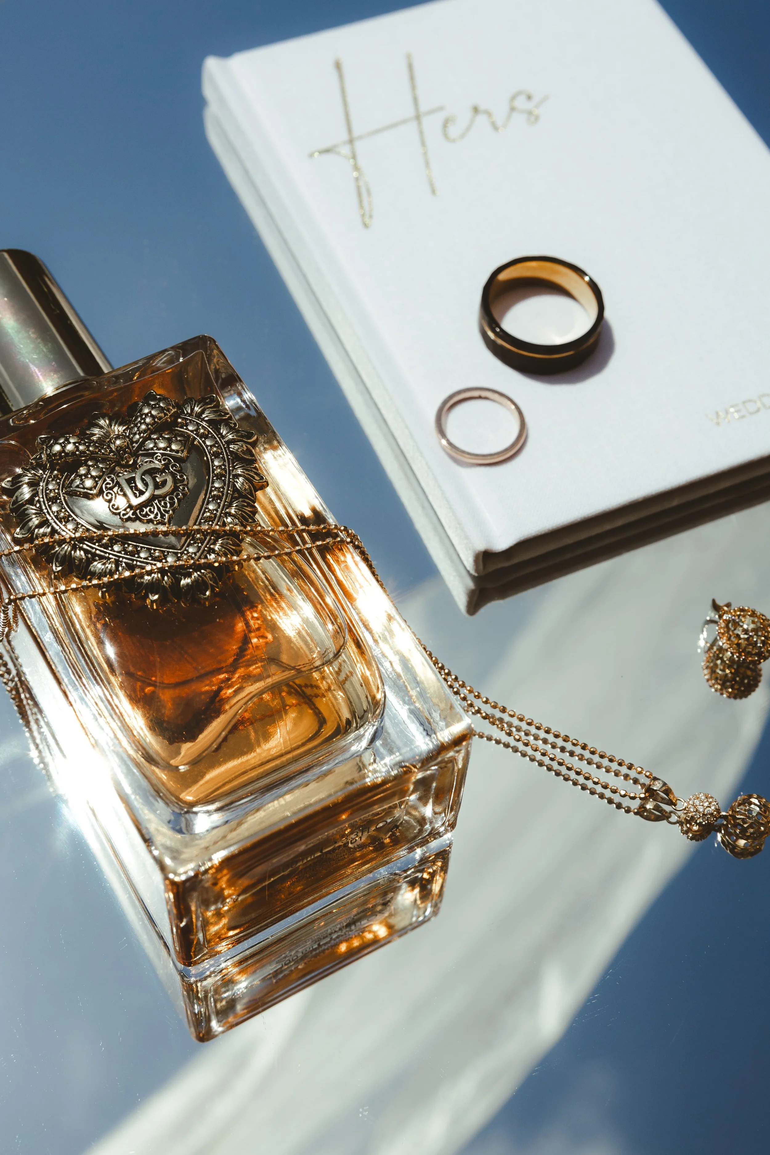 Close-up of a perfume bottle with gold jewelry on top, set on a reflective surface with a white book labeled 'Hers' in the background.