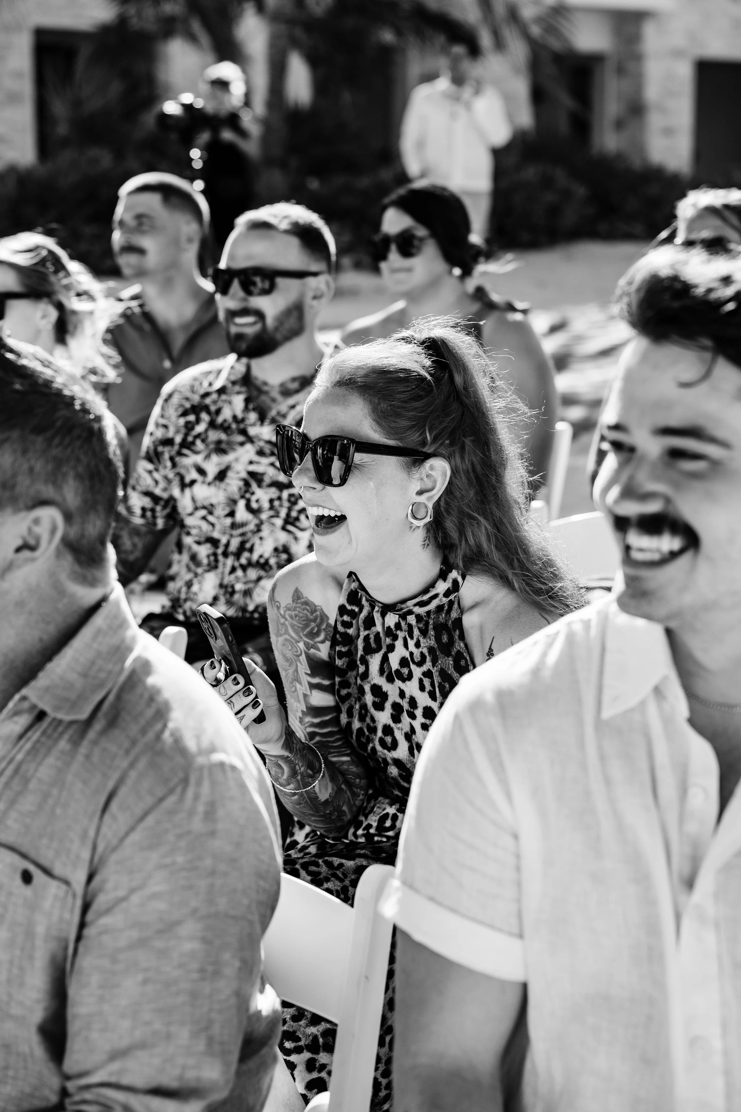 A group of people sitting in chairs outdoor, smiling, and engaging in conversation, with some wearing sunglasses. The background includes trees and a person standing in front of a building.