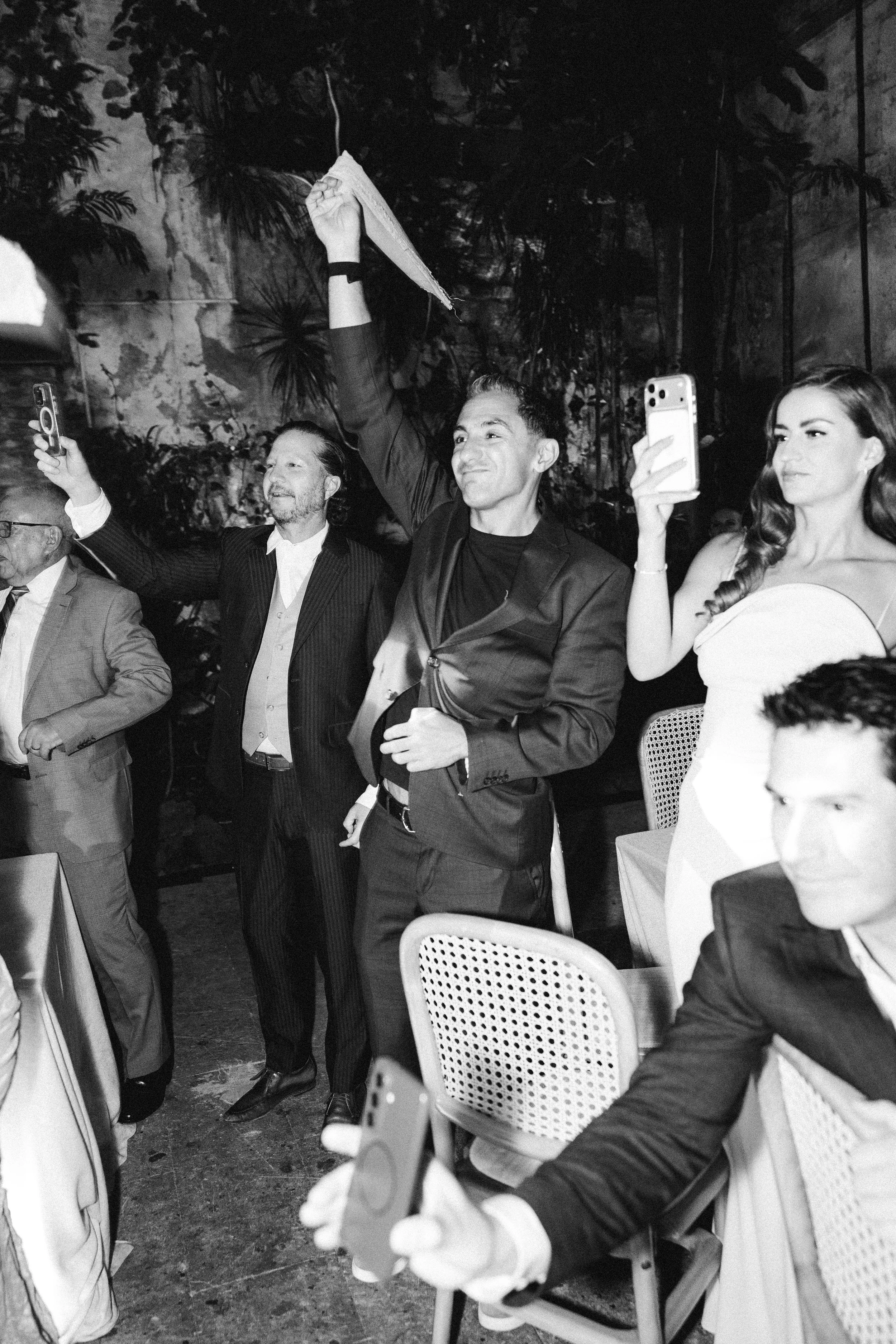 A black and white photo of a group of well-dressed people at an indoor event. One man holds a paper high, smiling, while a woman next to him takes a selfie. Others are engaging, with some seated and some standing, in a setting with chairs and plants.