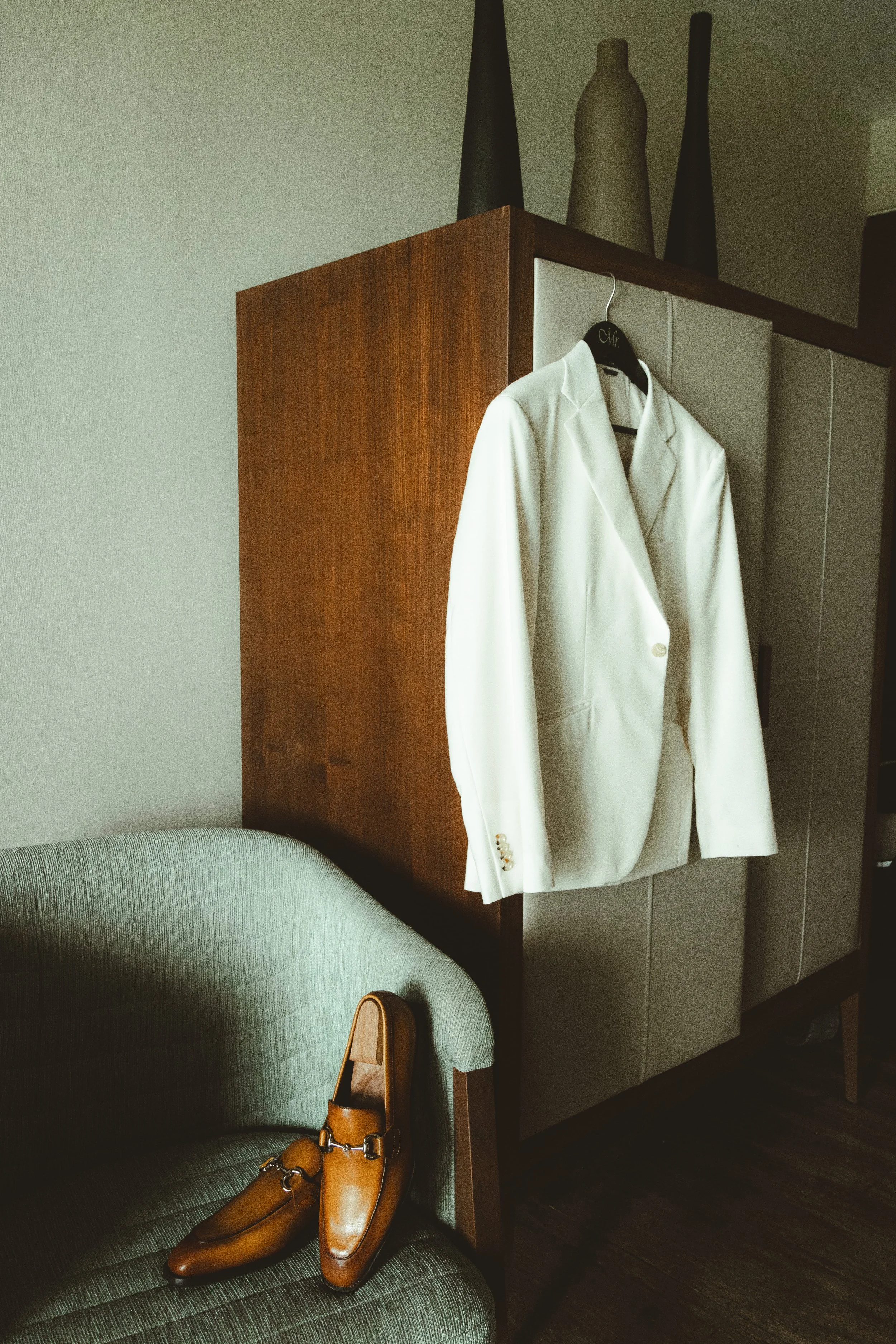A white suit jacket hanging on a black hanger labeled "Mr." on a wooden furniture piece. Brown dress shoes and a matching bag are placed on a green upholstered chair. Vases are seen on top of the furniture.
