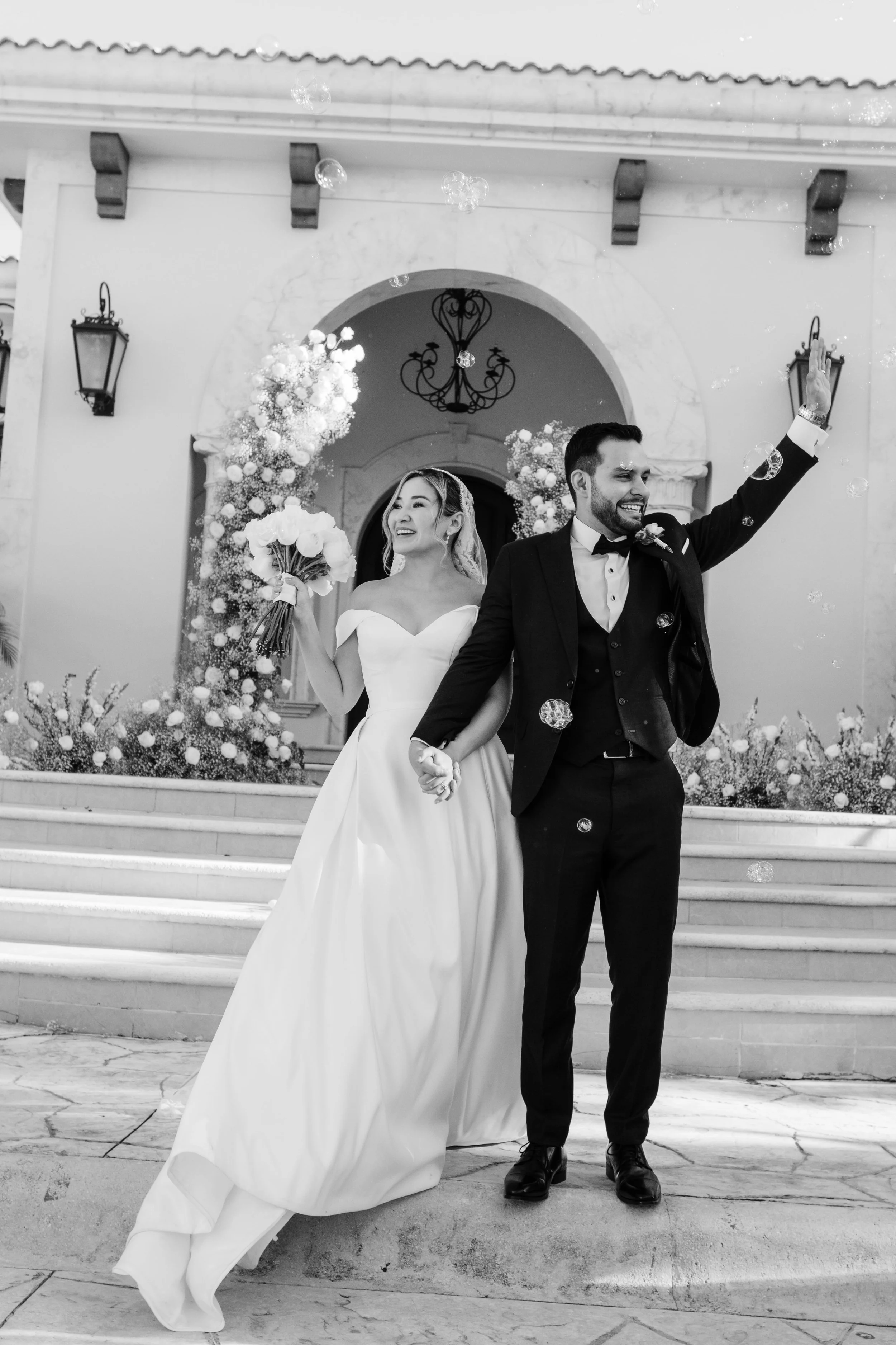 A bride and groom holding hands on staircase outside a building, the bride holding a bouquet, celebrating wedding, black and white photograph.