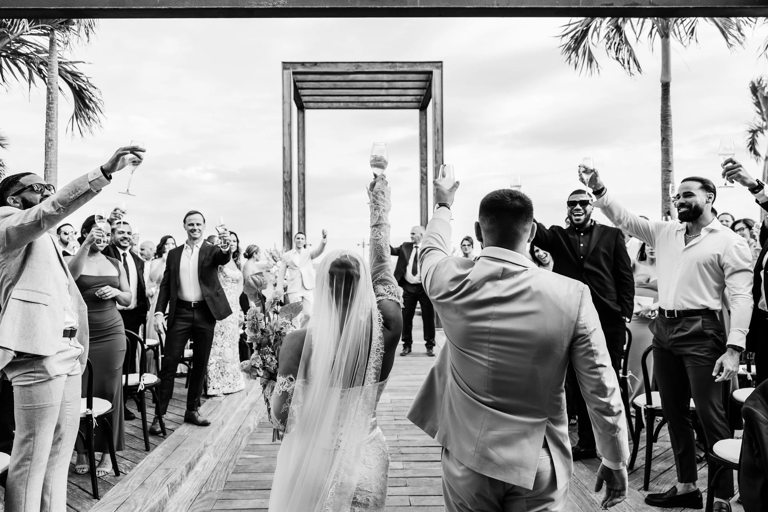 A black and white photo of a wedding celebration with guests raising glasses in a toast, outdoors near palm trees, with a bride and groom in the background.