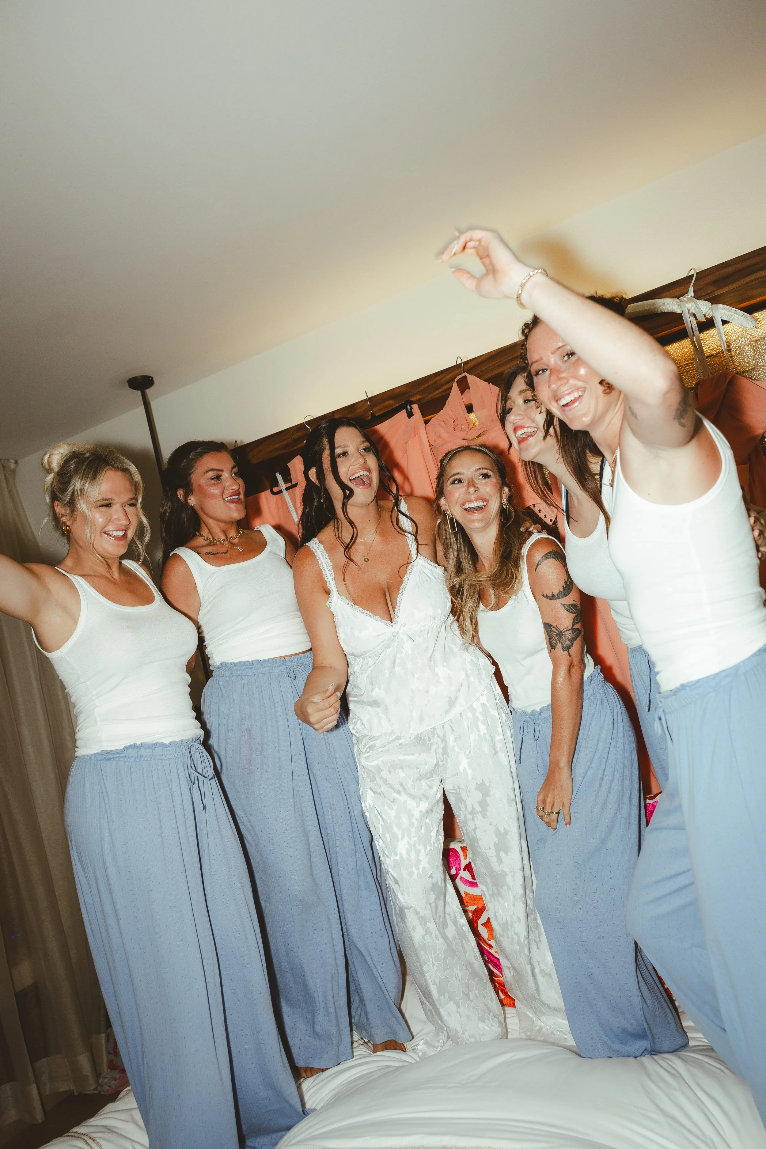 A group of women in pajamas laughing and celebrating on a bed, with their arms raised, in a cozy bedroom setting.