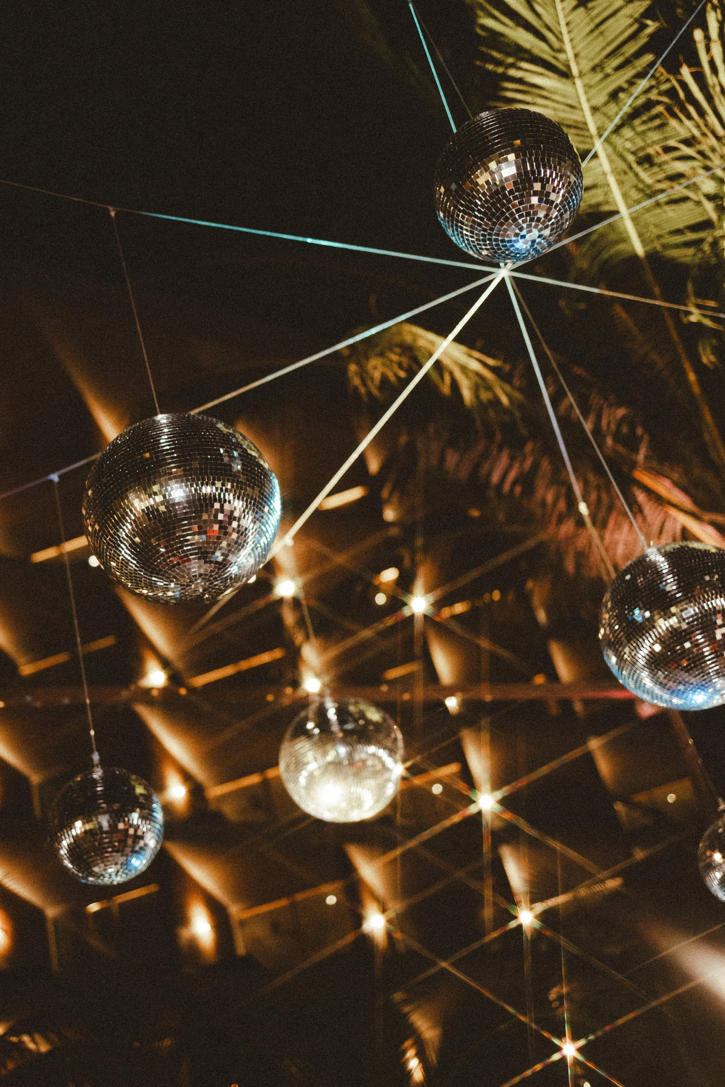 Multiple disco balls hanging from the ceiling, reflecting light, with wooden ceilings and palm leaves in the background. Wedding reception set up.