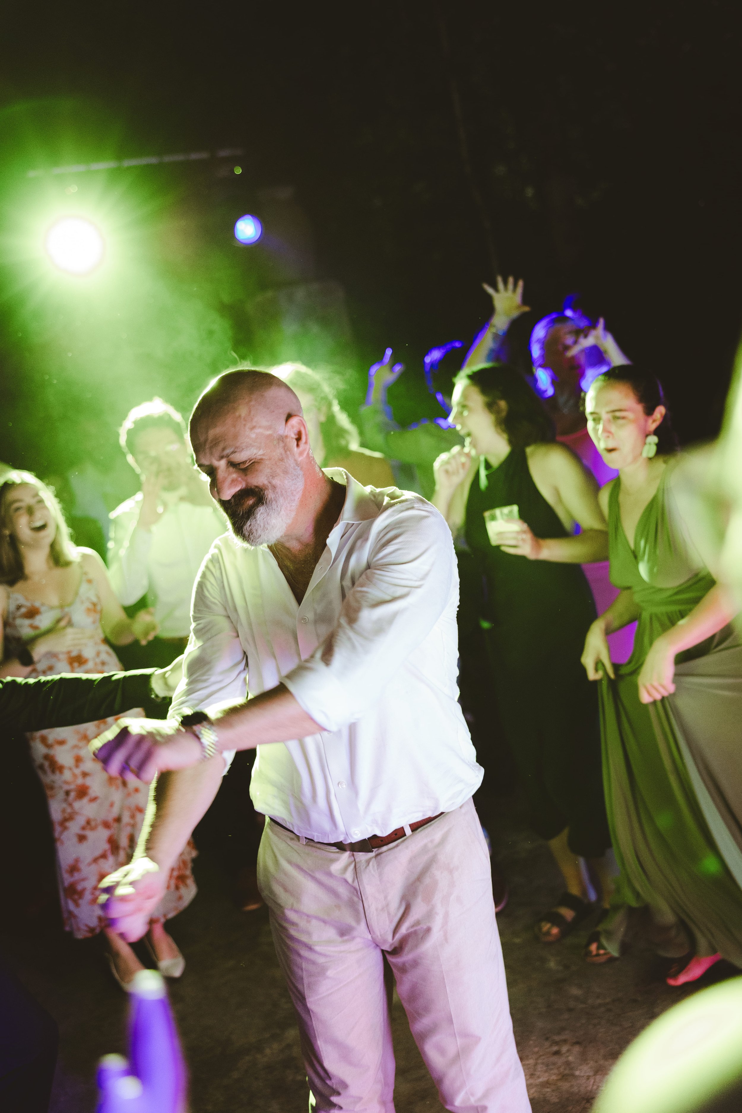 A man with a gray beard wearing a white shirt dancing at a party with colorful lighting, surrounded by women in dresses enjoying music and dancing.