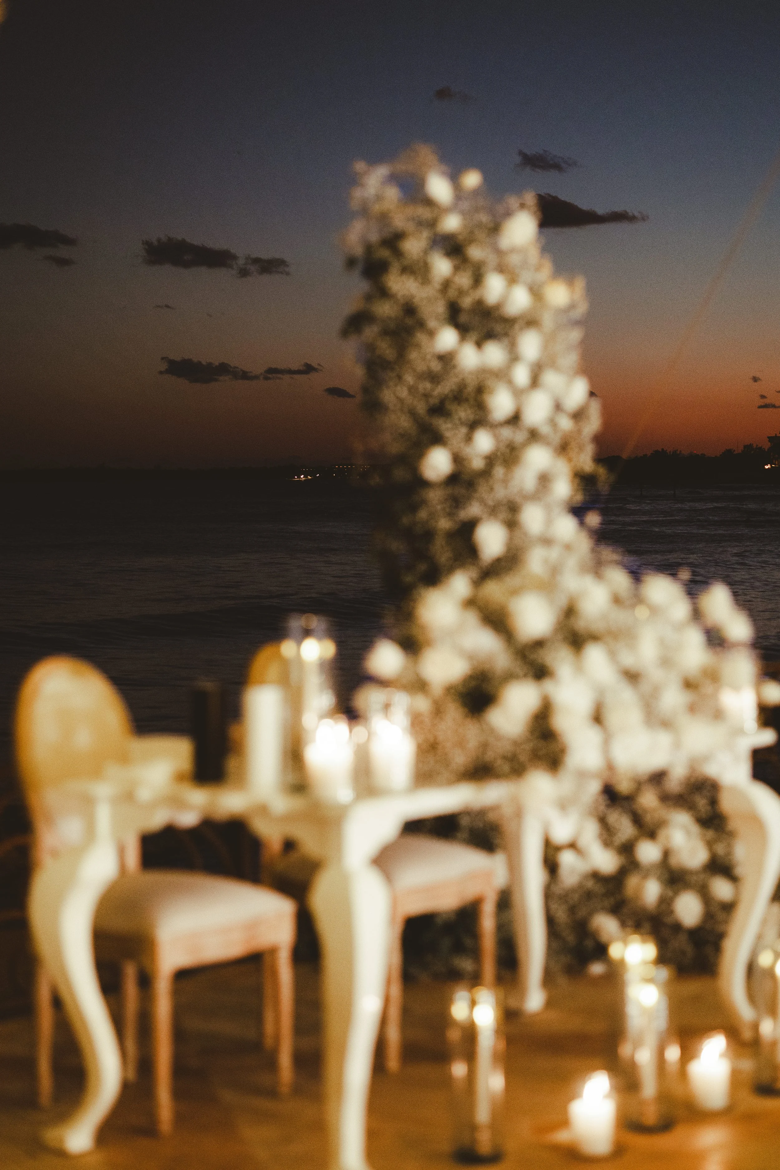 Indoor event setup with wooden chairs and white flowers, candles on the floor, overlooking a body of water during sunset or dusk, with a cloudy sky.