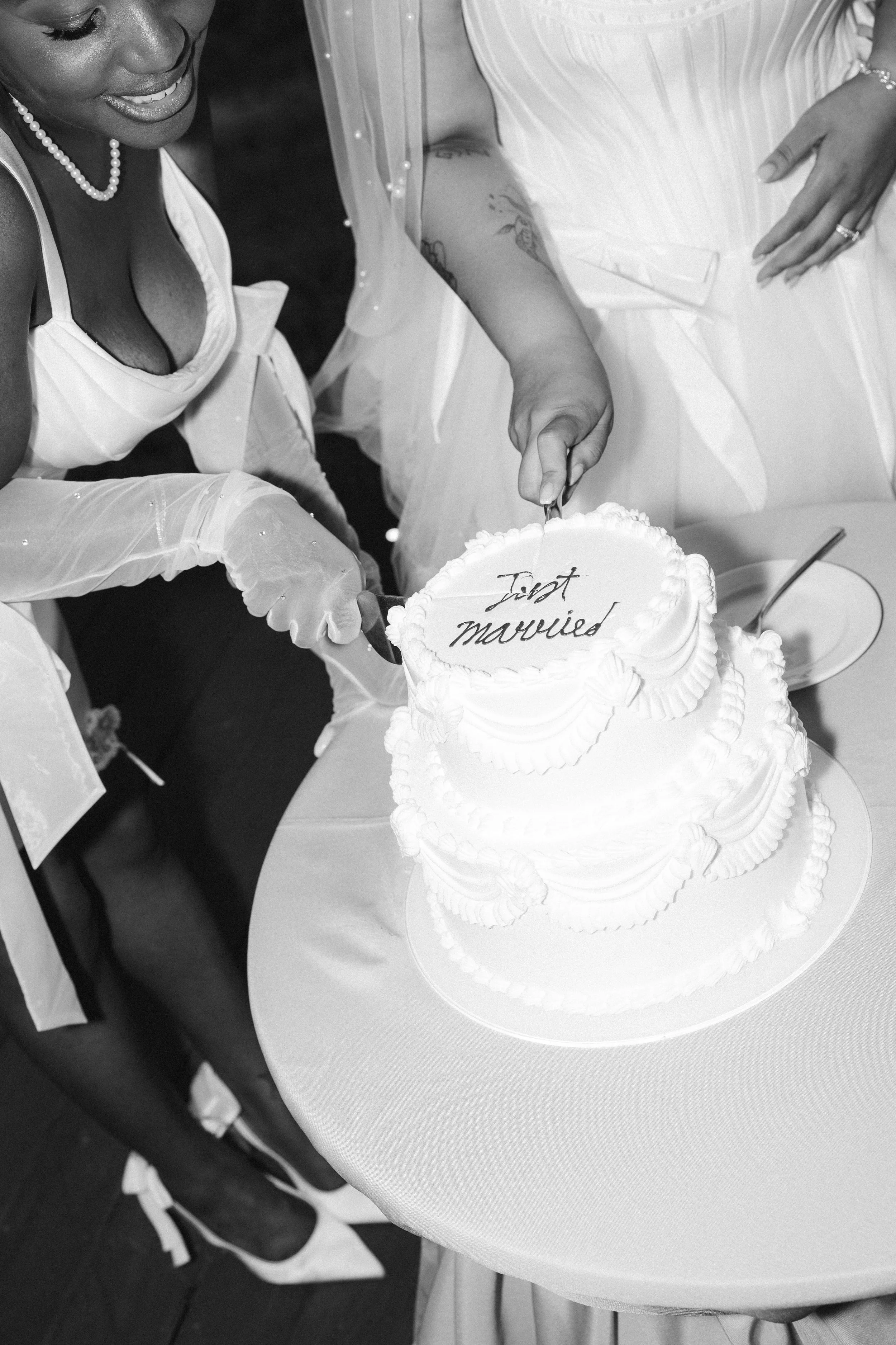 A black-and-white photograph of a woman in a wedding dress smiling while cutting a wedding cake. The cake has the words 'Just married' written on it.