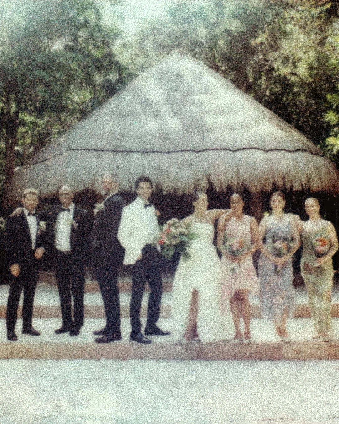 Group of people at a wedding photo, standing in front of a thatched roof hut surrounded by trees, with the bride, groom, and attendants dressed in formal attire and holding bouquets.