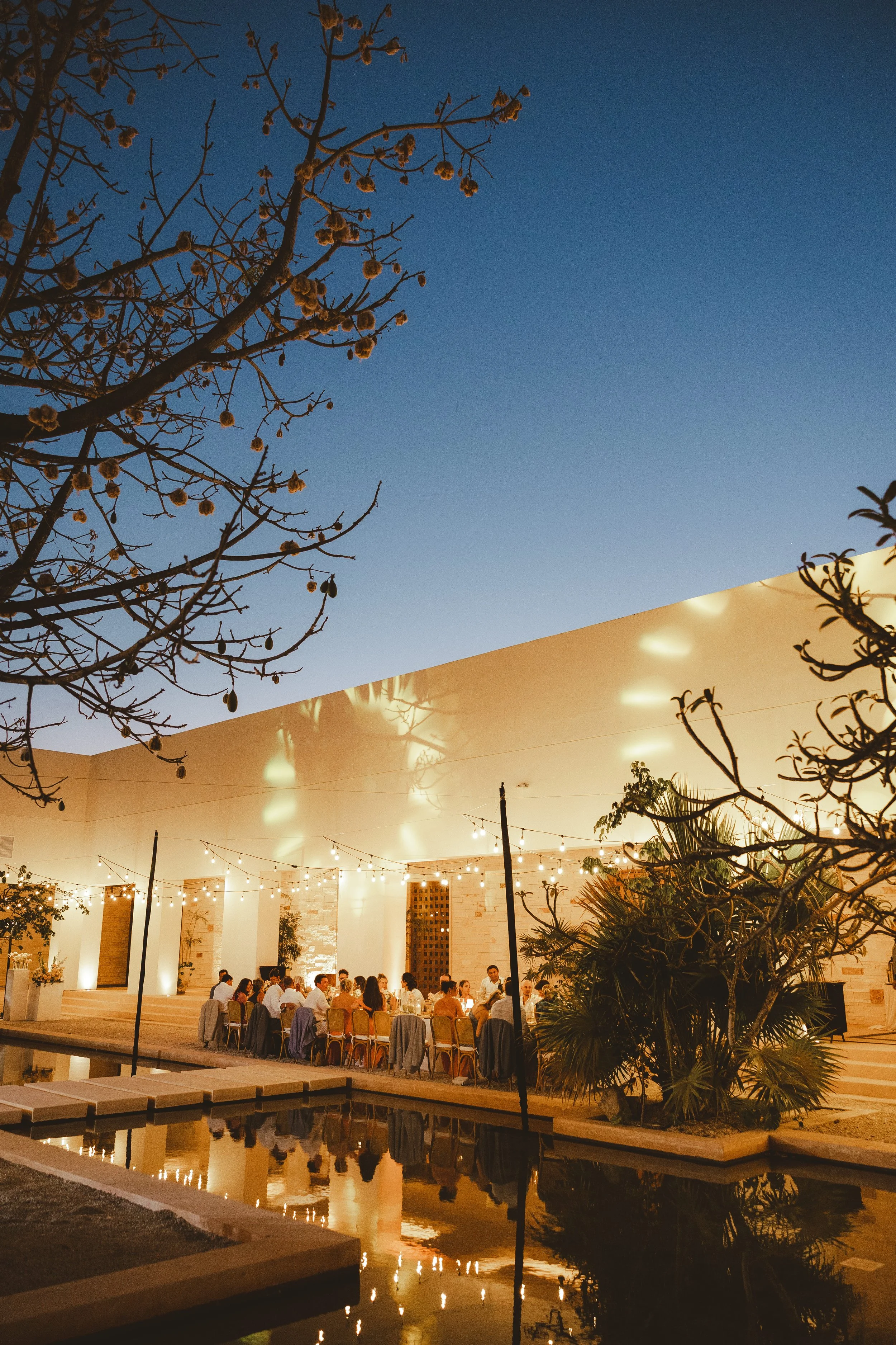 People dining outdoors during evening with string lights, a swimming pool, and trees against a twilight sky.