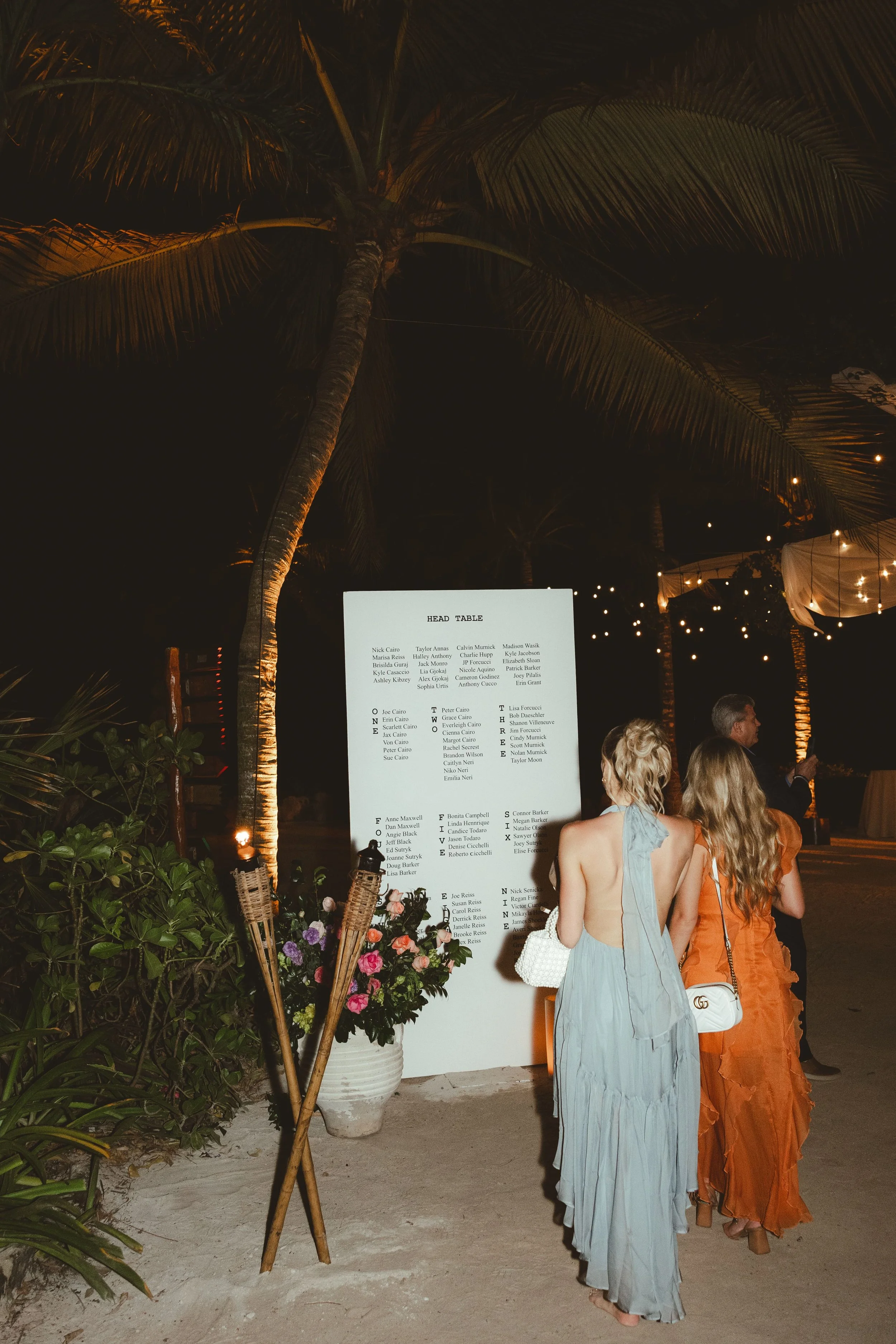 People dressed in elegant evening gowns standing near a large white seating chart at an outdoor event at night. There are string lights, plants, and a tall palm tree in the background.