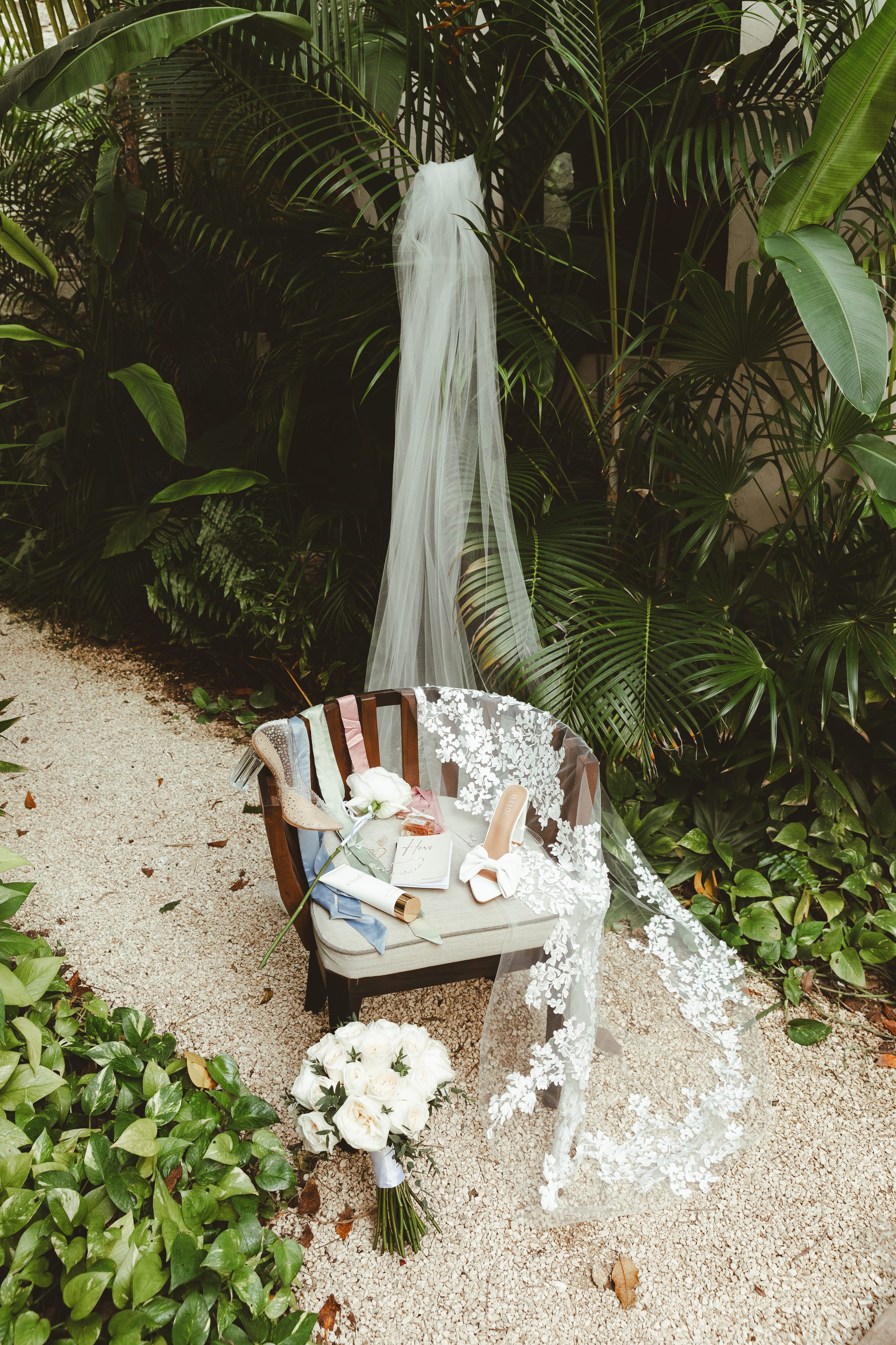A wedding setup with a small beige bench decorated with a white lace veil, a bouquet of white roses, and various small gifts and cards. The background features lush green tropical plants, and a sheer white veil is hanging from a plant.