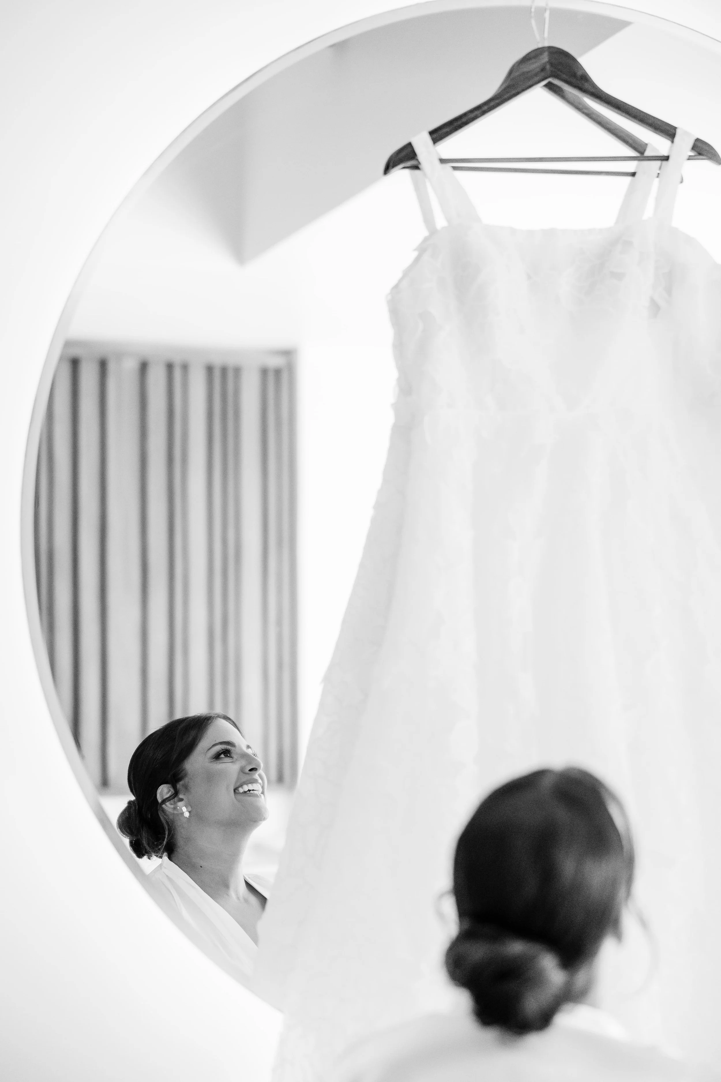 A woman smiling and looking up at a wedding dress hanging on a hanger in front of a mirror.