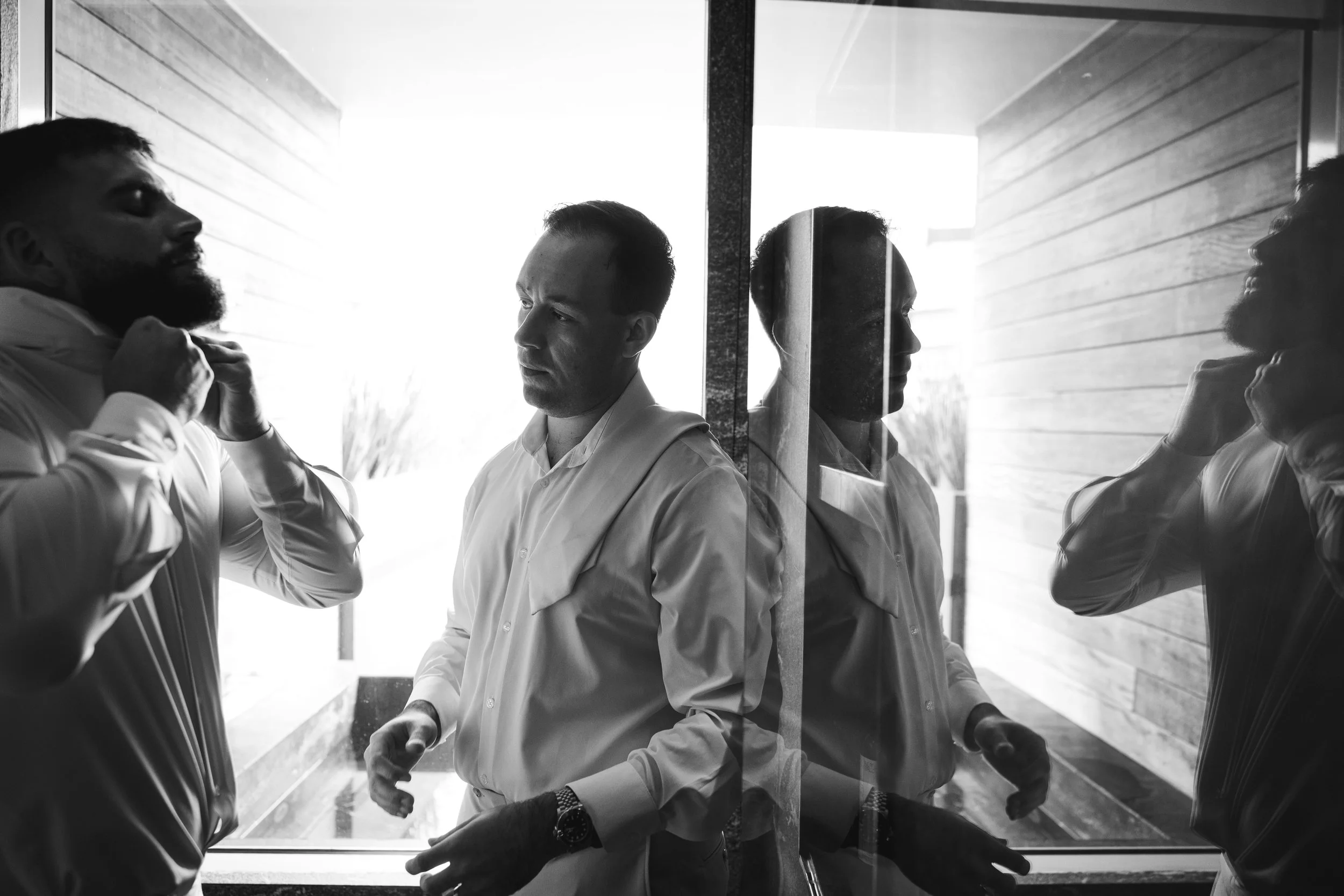 Black and white photo of three men standing near a large window, with two of them looking in opposite directions and the middle man looking ahead, reflection visible on glass.