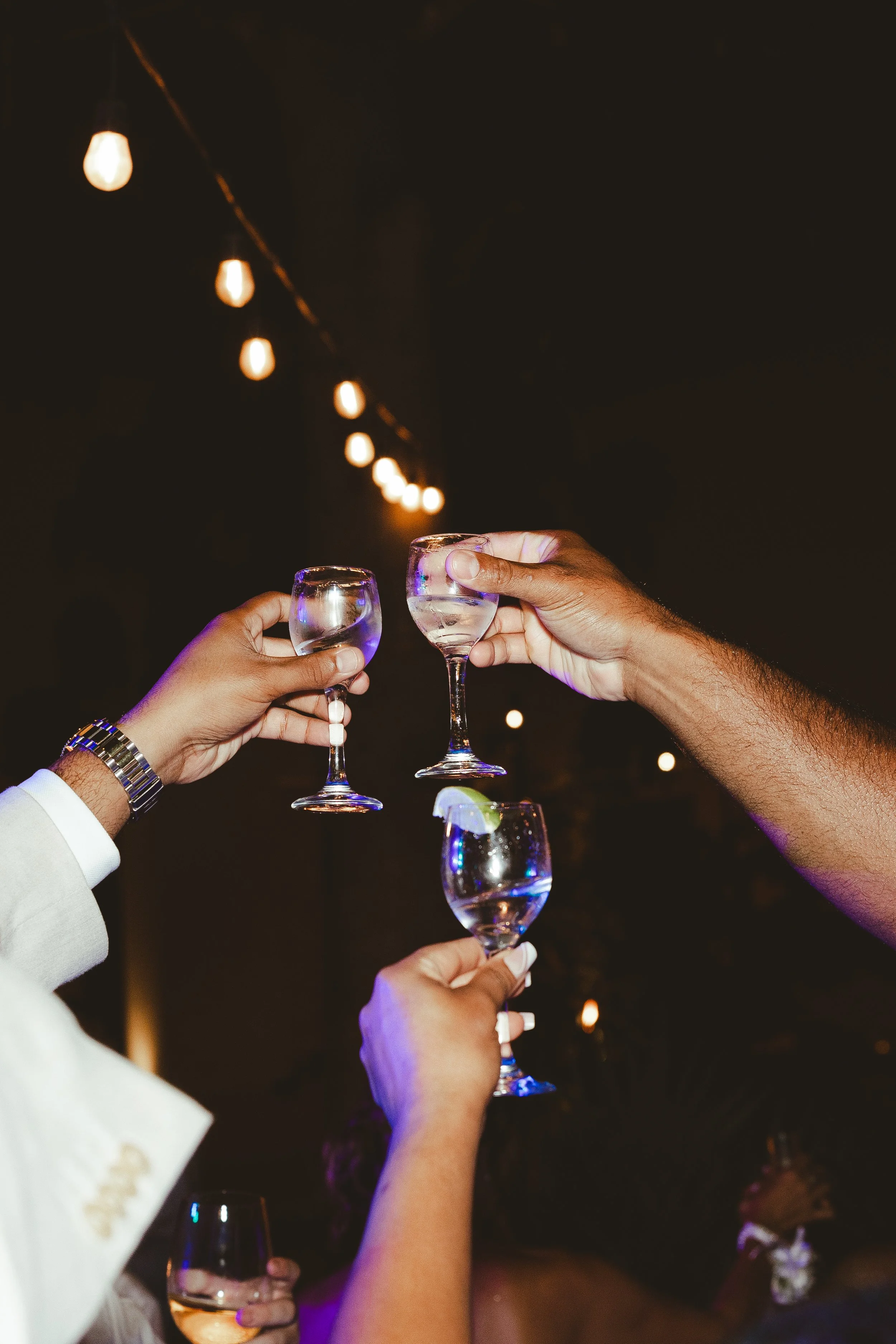 People clinking glasses of champagne in a toast at a celebration, nighttime setting with string lights overhead during wedding reception party.