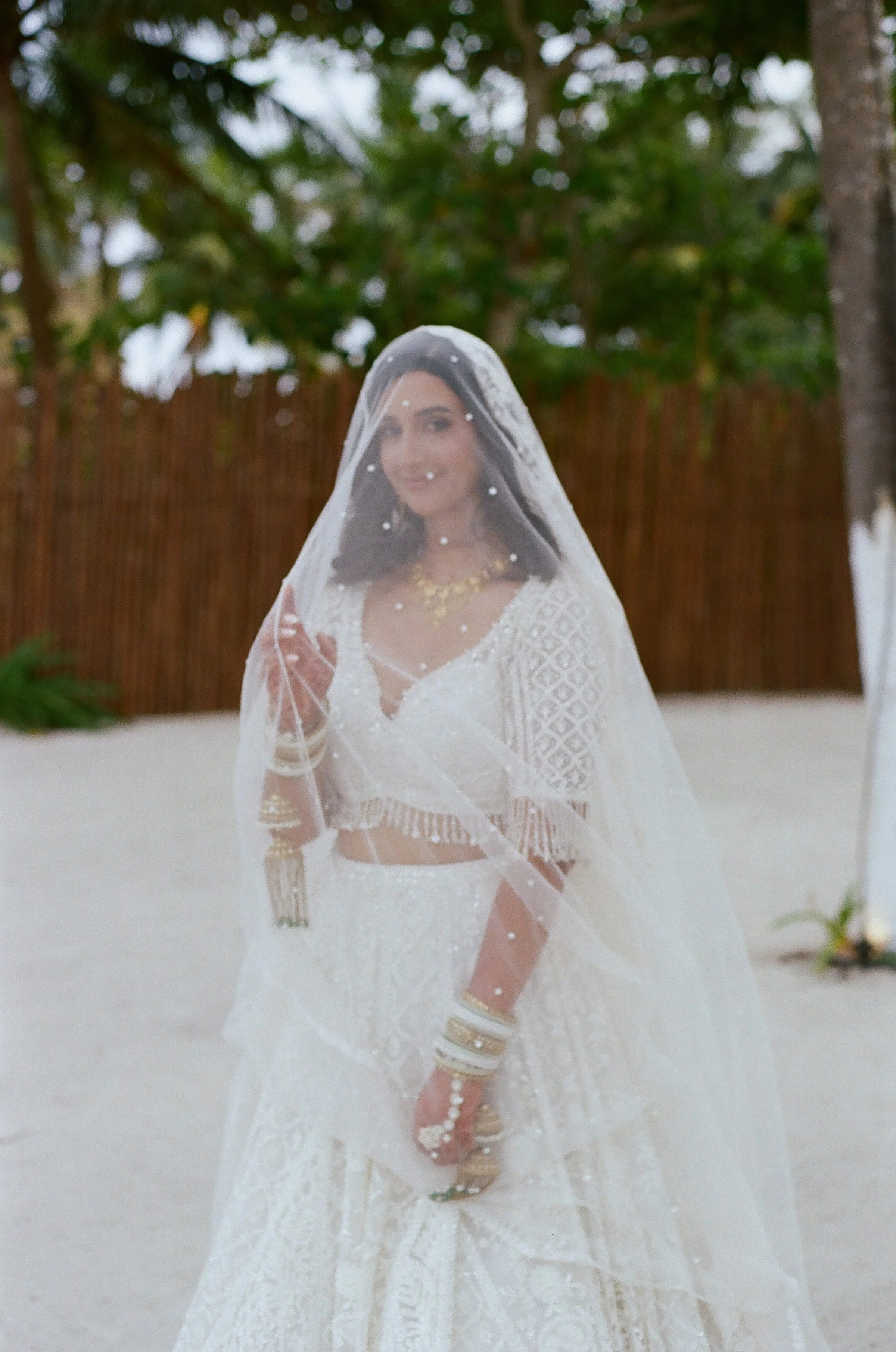 A bride dressed in a white traditional indian outfit with gold jewelry, standing outdoors on a sandy area with trees and a wooden fence in the background, wearing a sheer veil over her head and smiling.