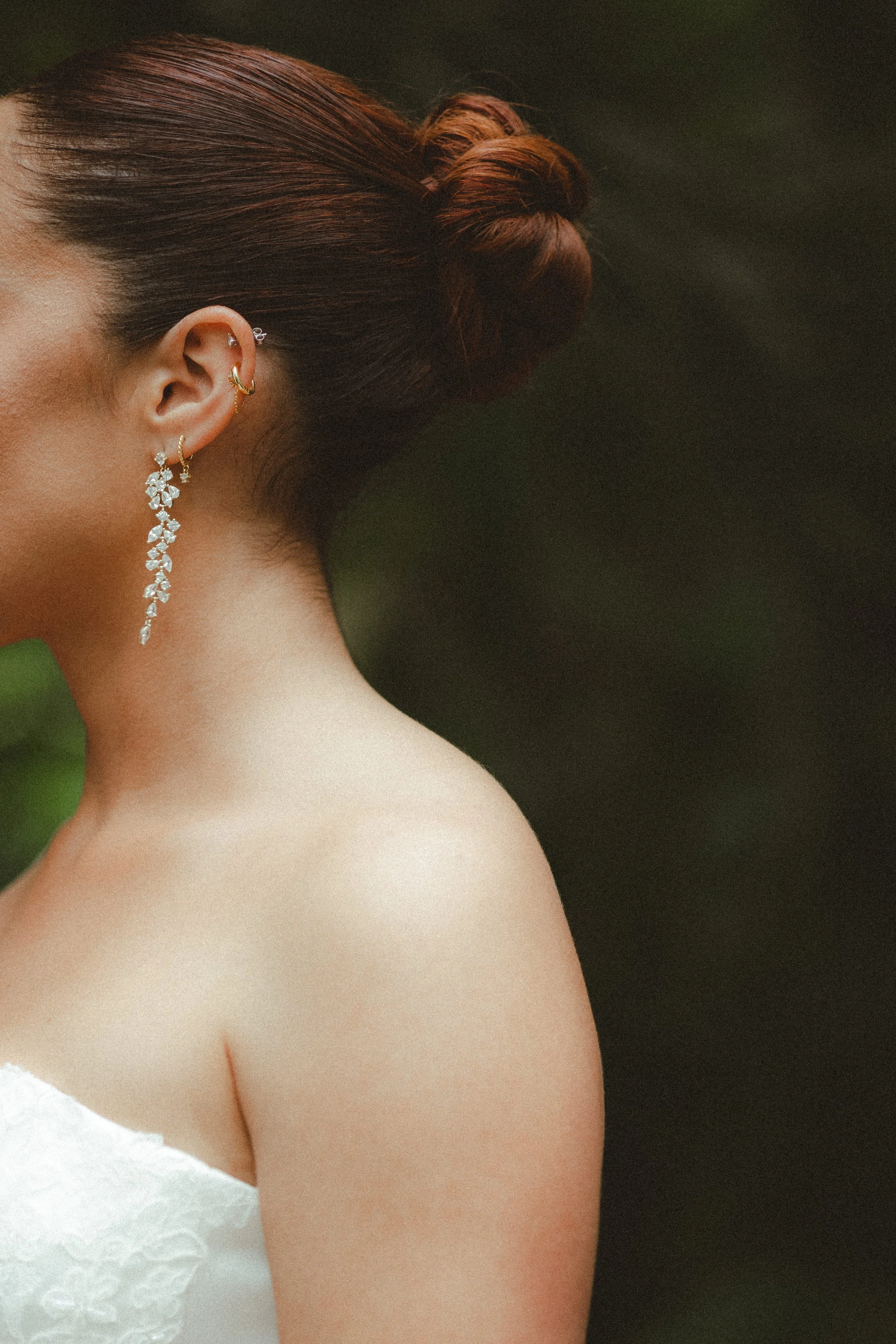 Close-up of a bride with red hair styled in a bun, wearing multiple earrings including a long, dangling chandelier earring and a small hoop, with a white lacy strap visible on her shoulder against a dark background.