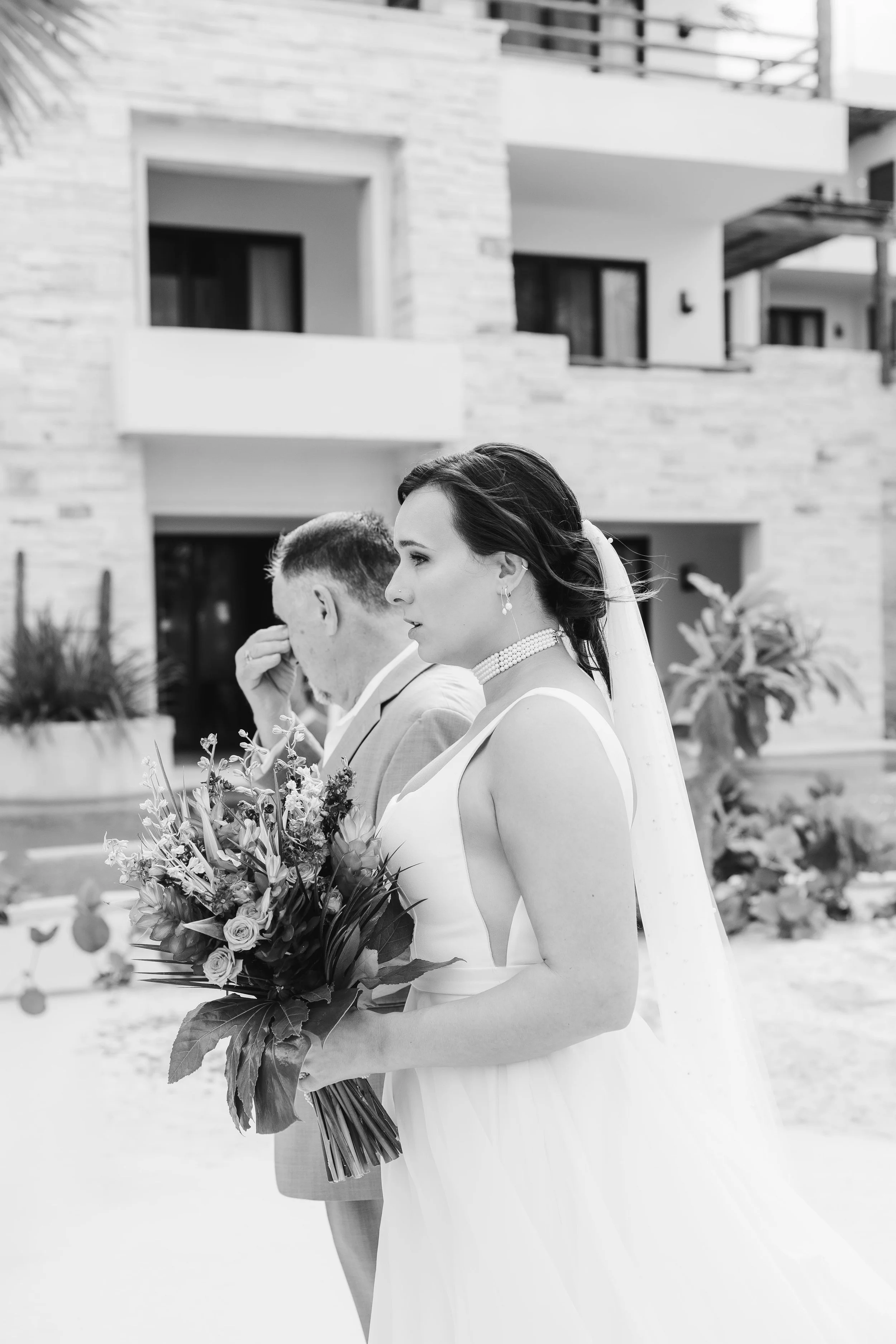 Black and white photo of a bride holding a bouquet, standing outdoors with modern residential buildings in the background.