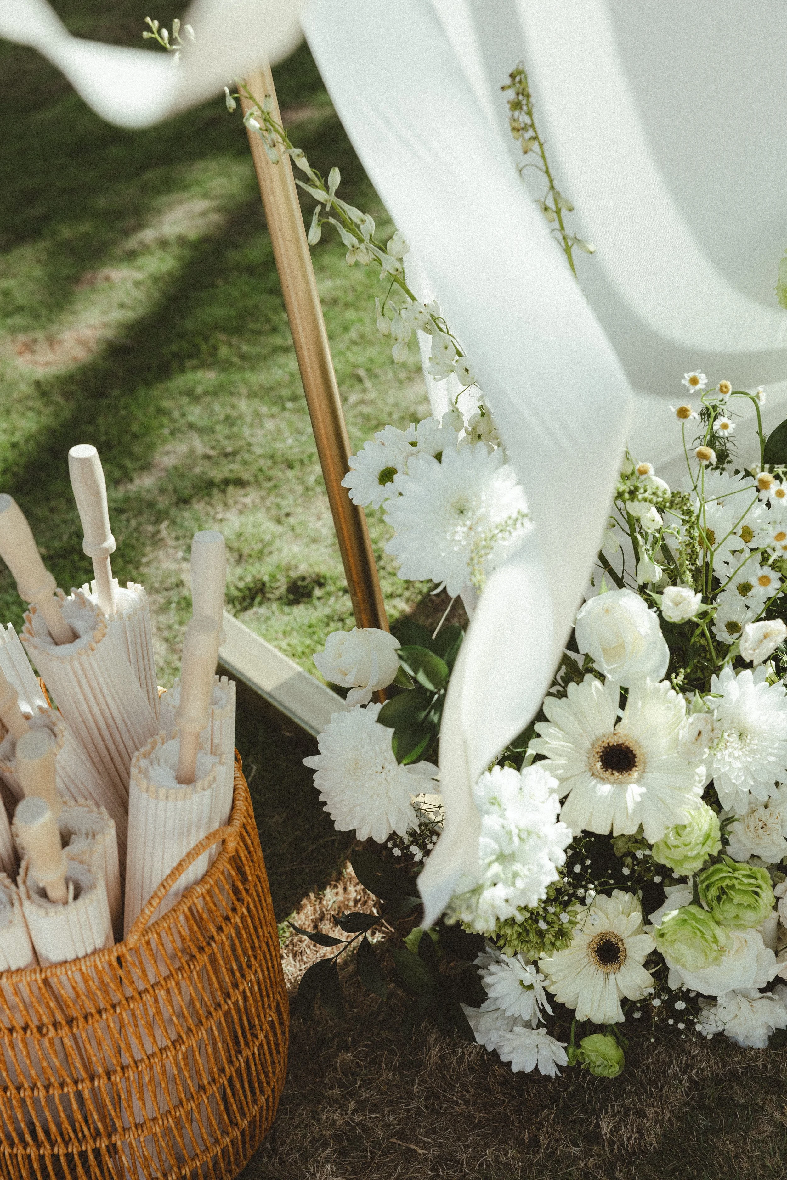Close-up of a floral arrangement with white flowers and greenery, with a wicker basket holding paper umbrellas in the foreground.