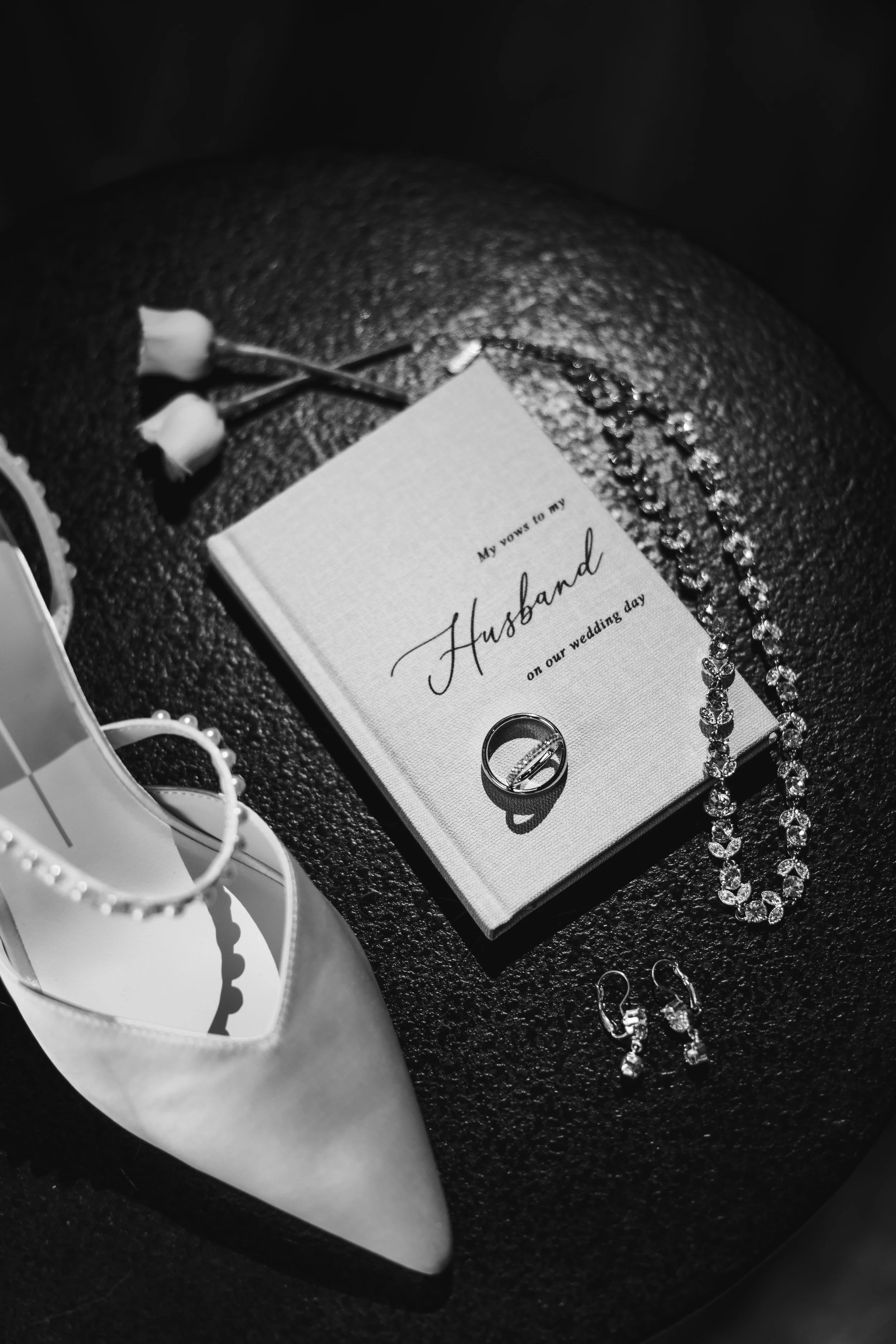 Black and white photo of wedding accessories including a small book titled "My vows to my husband on our wedding day", jewelry such as a rhinestone necklace, rings, earrings, and a boutonniere with two white flowers. A pair of white shoes with a rhin