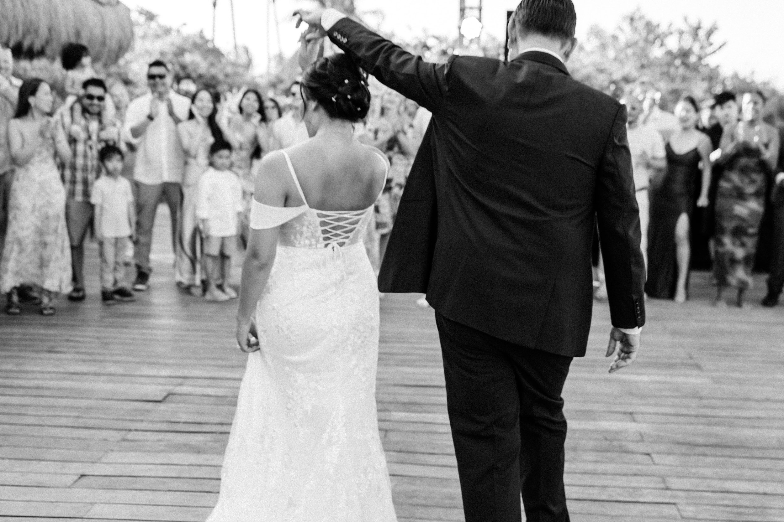 A bride and groom dancing at their wedding reception, with guests watching in the background on a wooden floor outdoors.