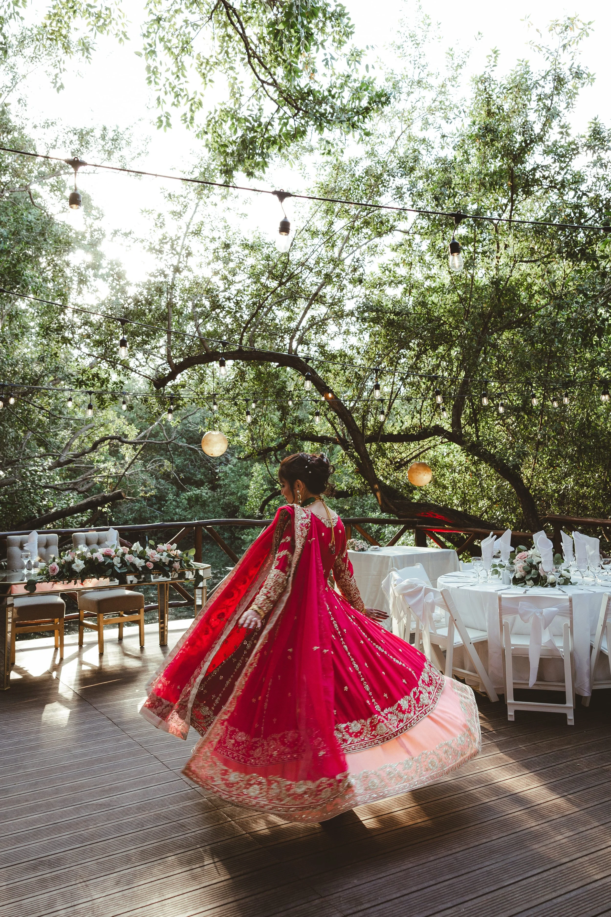 A woman in a red and gold traditional Indian dress, resembling a bridal outfit, is dancing outdoors under string lights and trees. There are white tables and chairs with floral centerpieces in the background, indicating a festive event or celebration