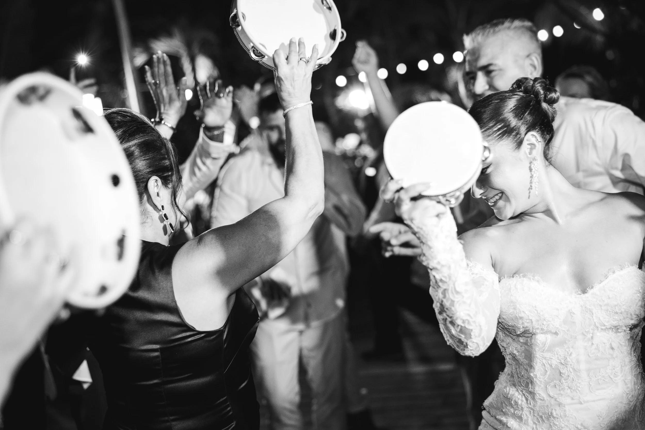 People dancing at a celebration, with some holding tambourines, in an outdoor nighttime setting.