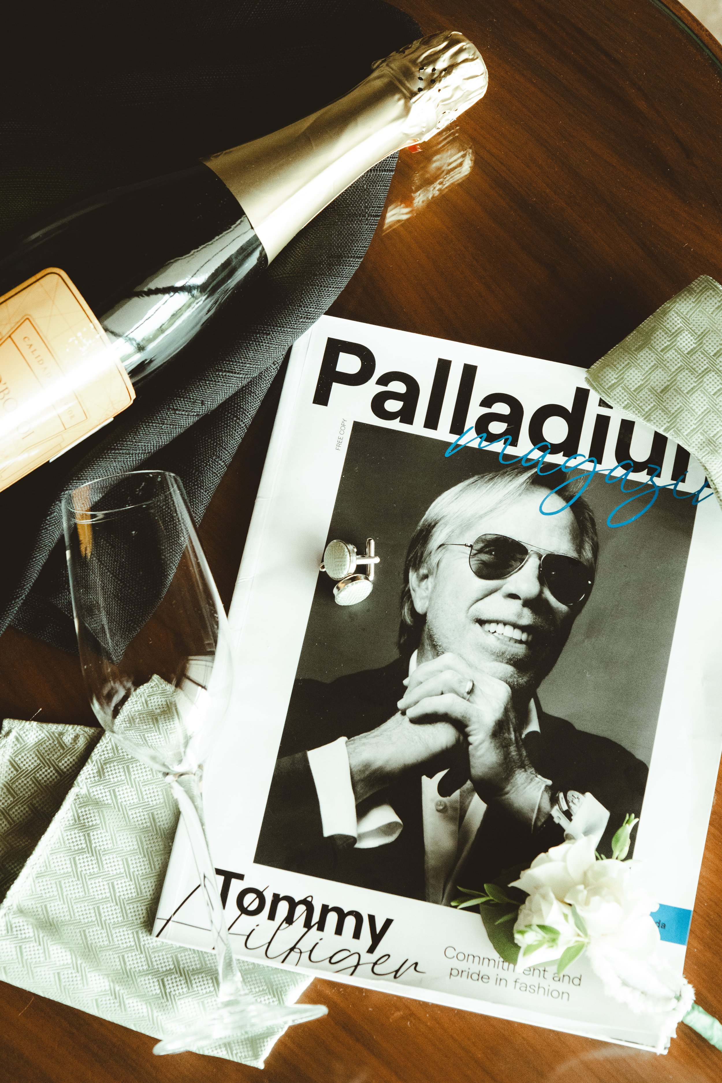 A magazine featuring a black and white photo of an older man smiling and wearing sunglasses. The magazine is titled "Tommy" and has the word "Influencer" in script. Next to the magazine is a bouquet of white flowers and a small green card, all placed