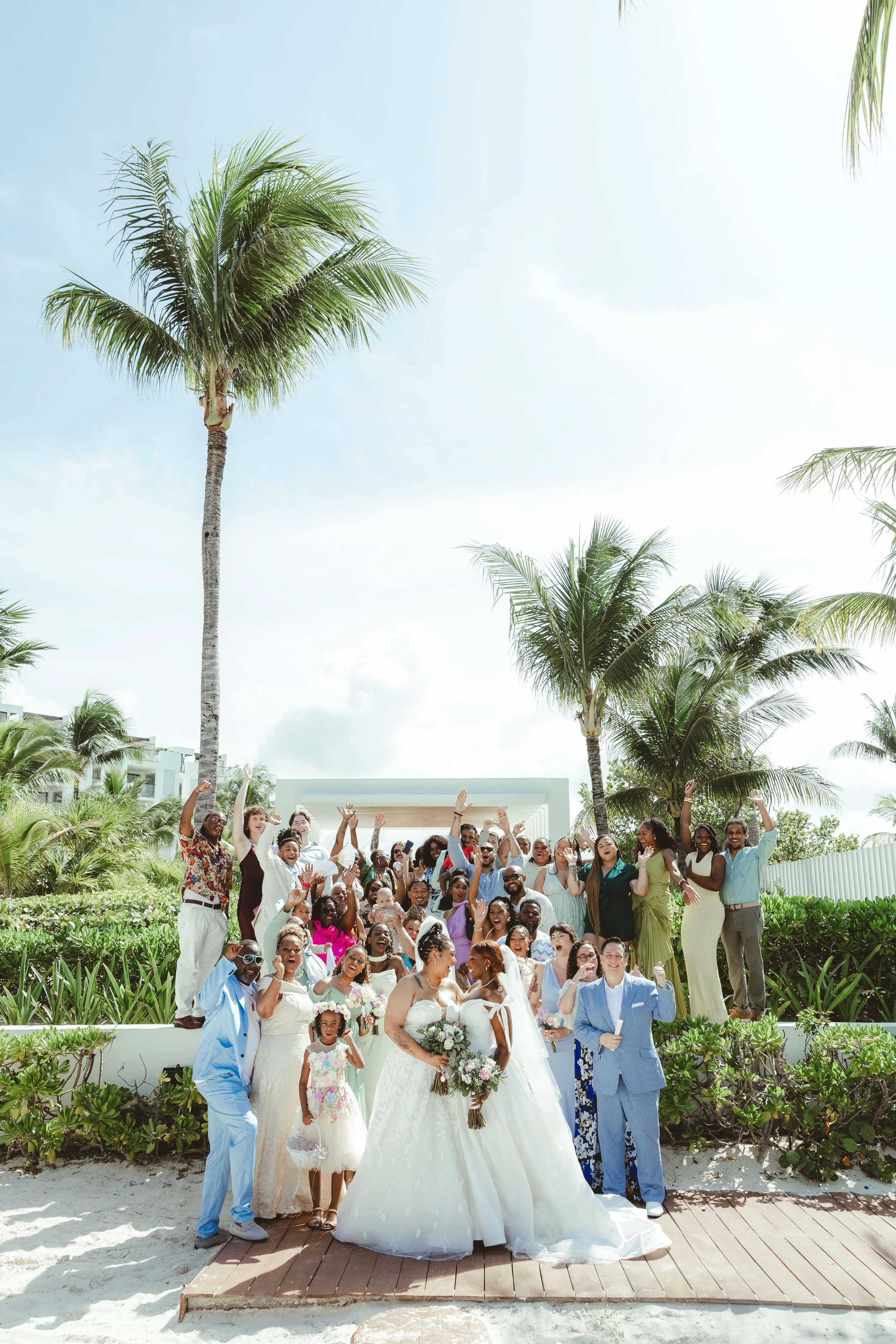 A large group of diverse people celebrating a wedding on a beachside garden. The bride and groom are in front, surrounded by friends and family, with palm trees and blue sky in the background.