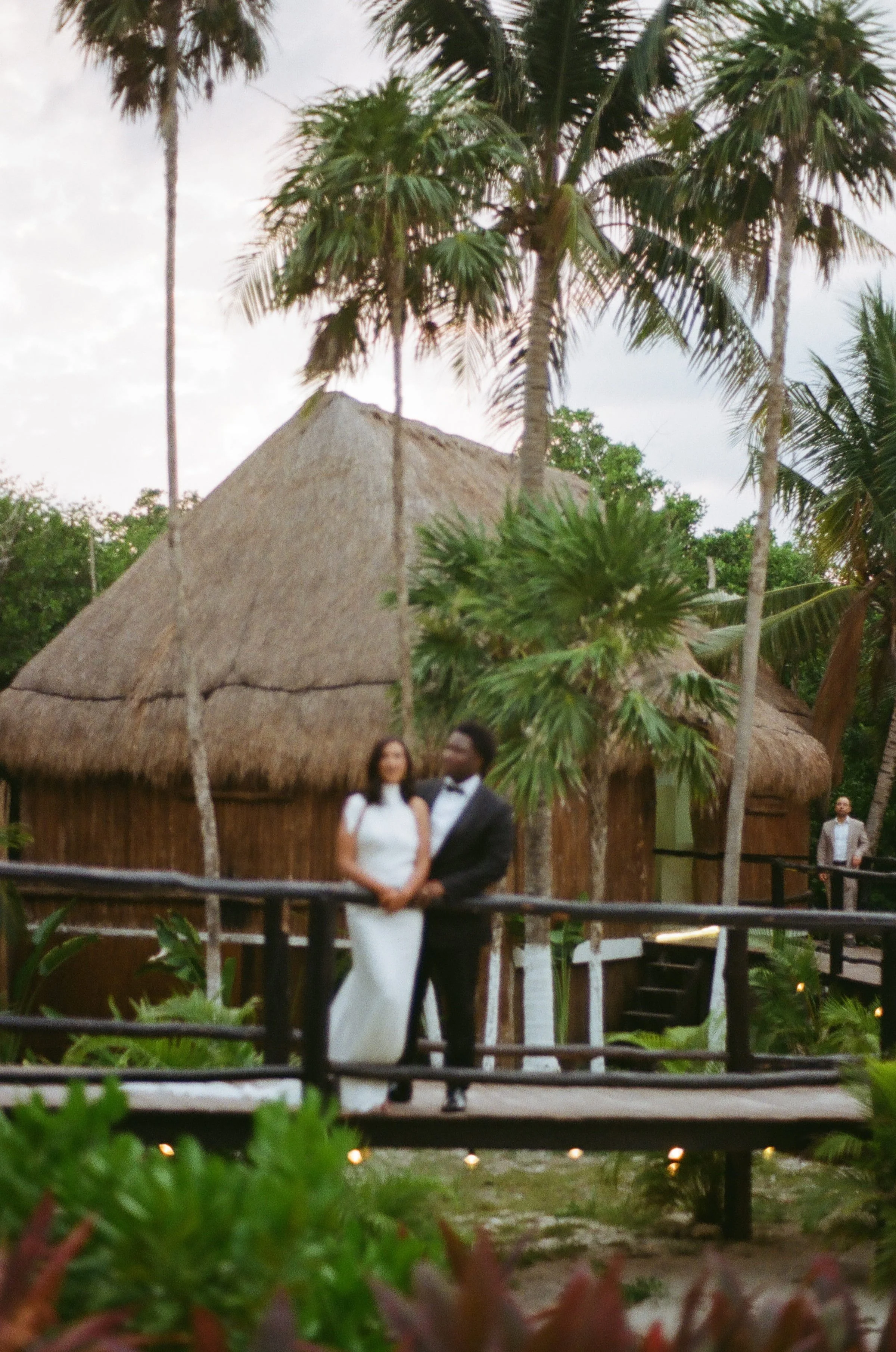 A couple, a woman in a white dress and a man in a tuxedo, stands together in front of a thatched-roof hut, with tropical trees and greenery around them, at Blue Venado Paamul Wedding Venue.