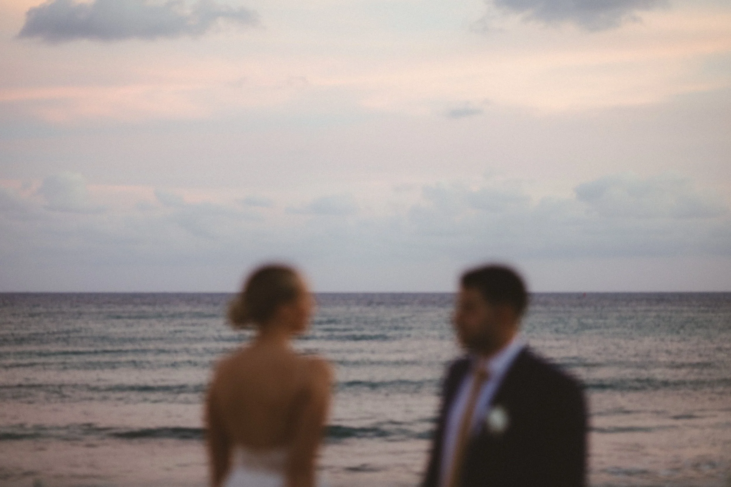 Two blurry people, a bride and a groom, standing on the beach near the water at sunset or dusk, with the ocean and cloudy sky in the background.