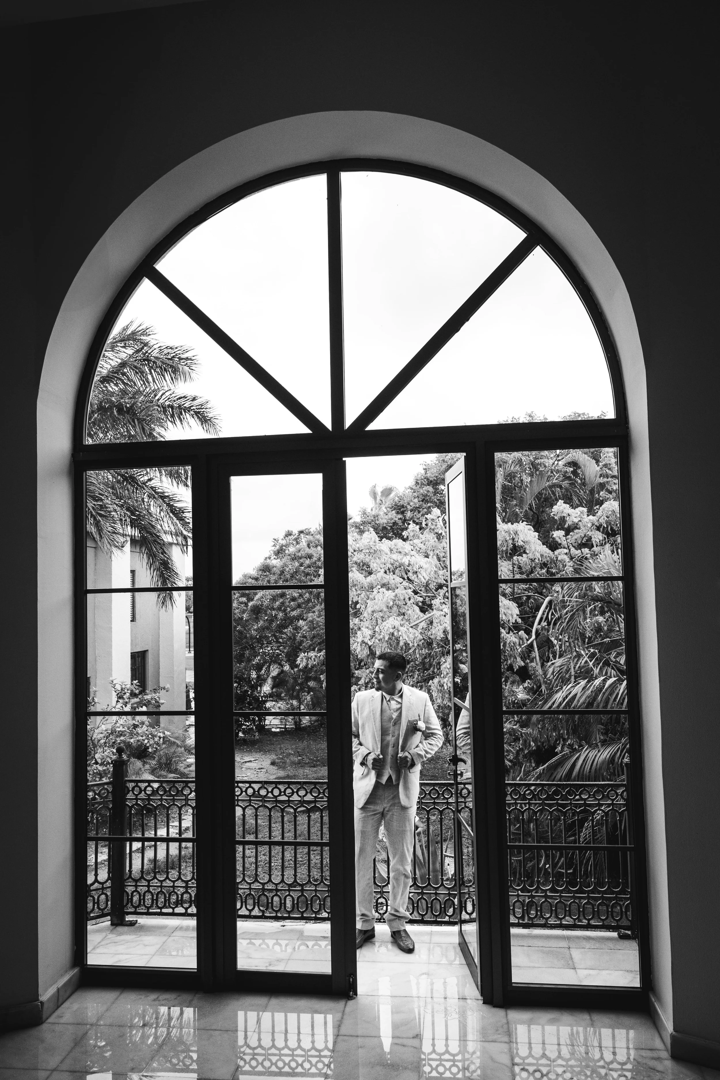 Black and white photo of a groom in a suit standing in a doorway, with a garden visible outside through a large arched window and glass doors in Iberostar Paraiso Lindo Spa.