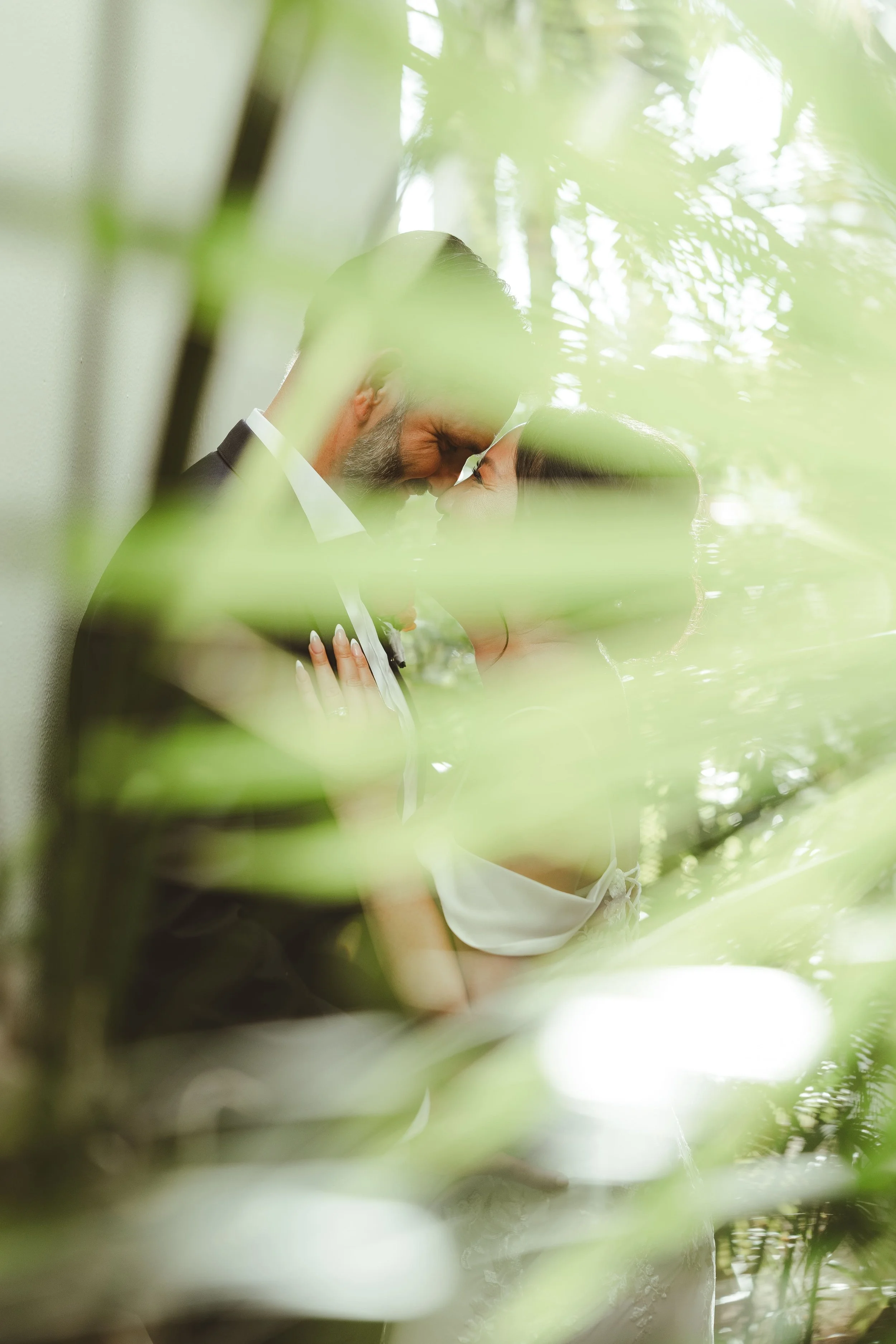 A couple is sharing a kiss, with their foreheads touching, amidst green foliage, and appears to be in a natural outdoor setting.