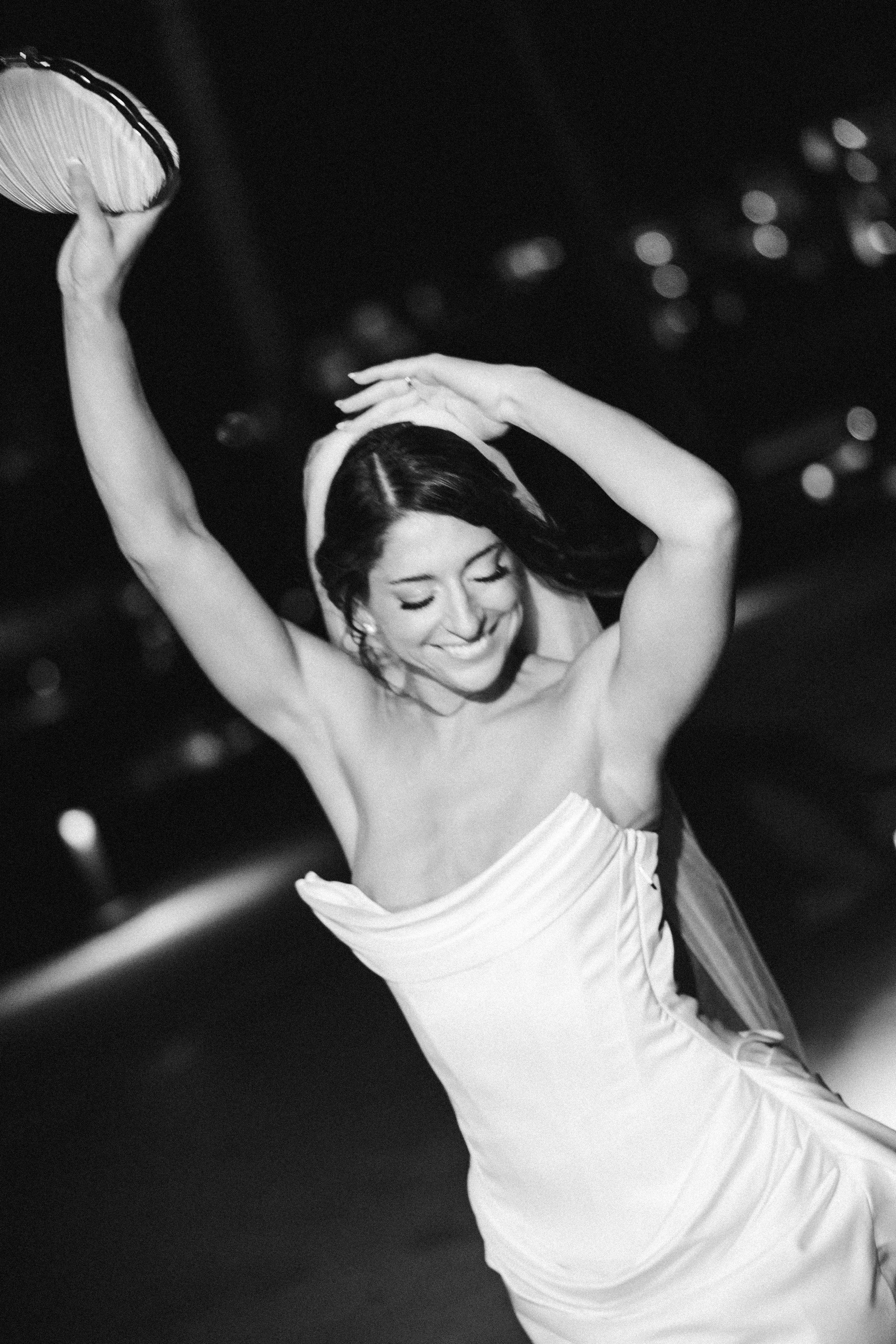 A woman with dark hair smiling, wearing a strapless dress, holding a parasol, and dancing or celebrating against a blurred dark background with bokeh lights.