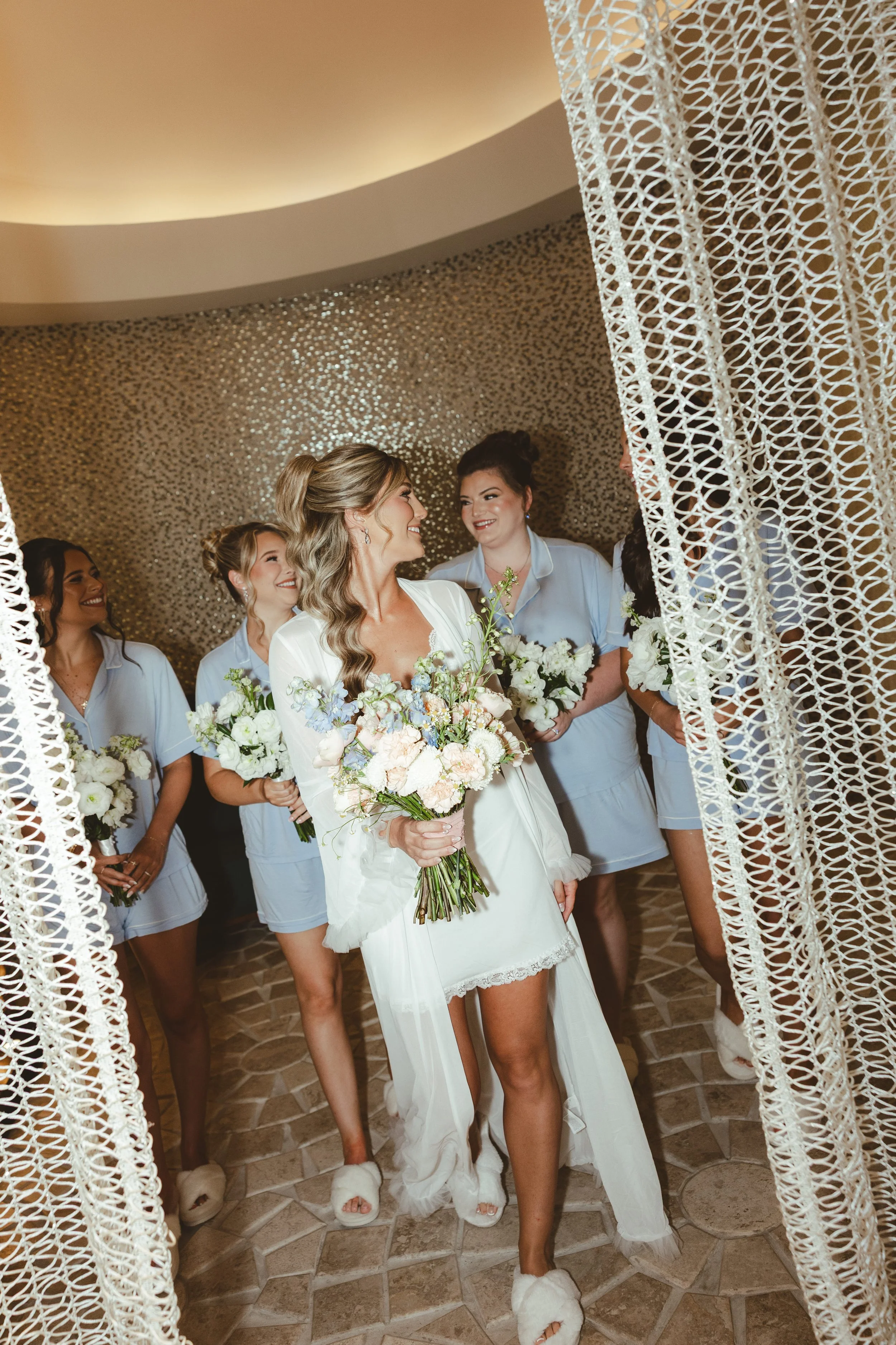 A bride with long, wavy hair smiles and walks through a lace curtain with her bridesmaids behind her in a wedding reception room with a glittery gold wall.