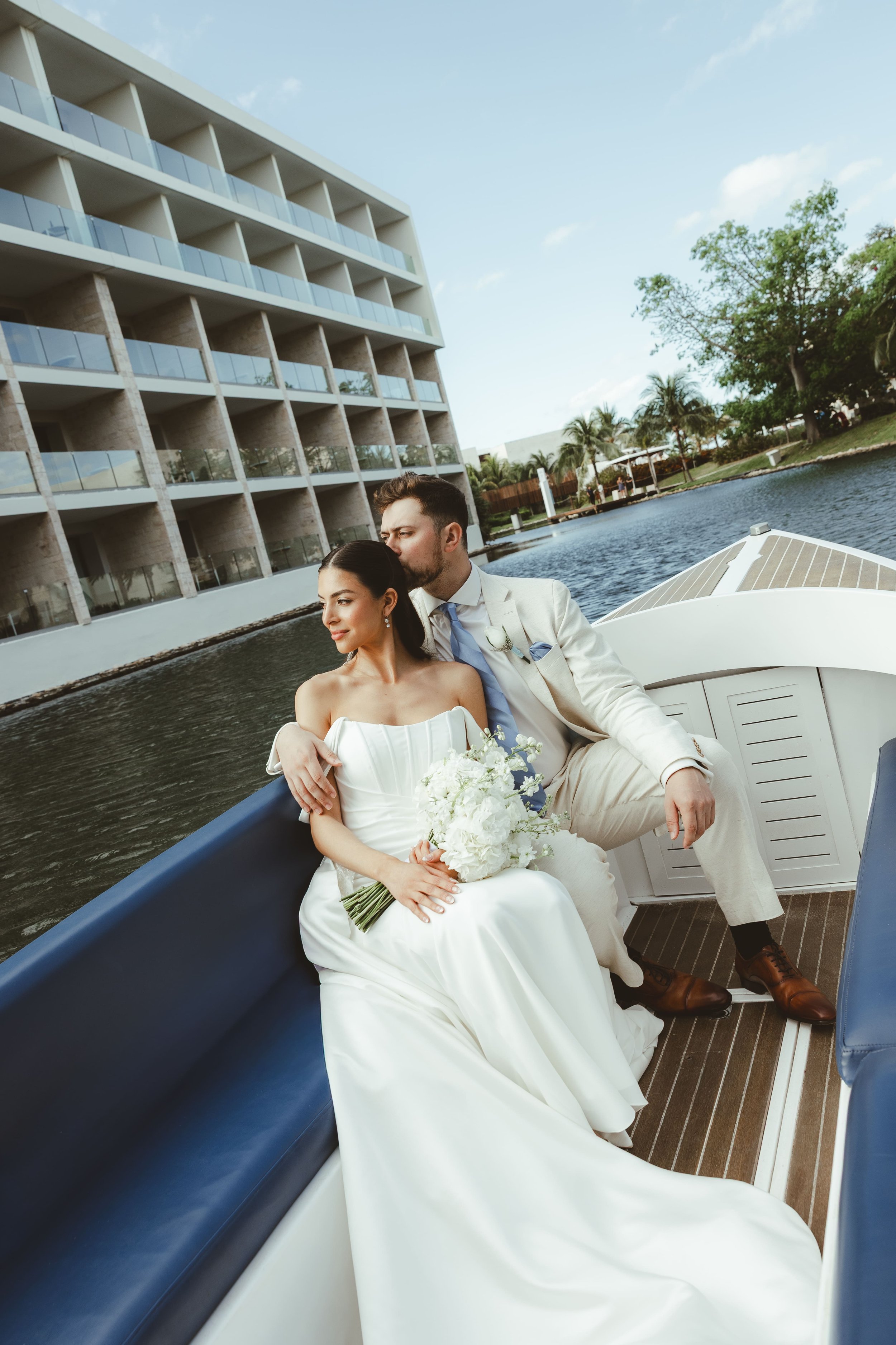 A newlywed couple on a boat, the bride holding a bouquet of white flowers and the groom kissing her temple, with a modern building and trees in the background.