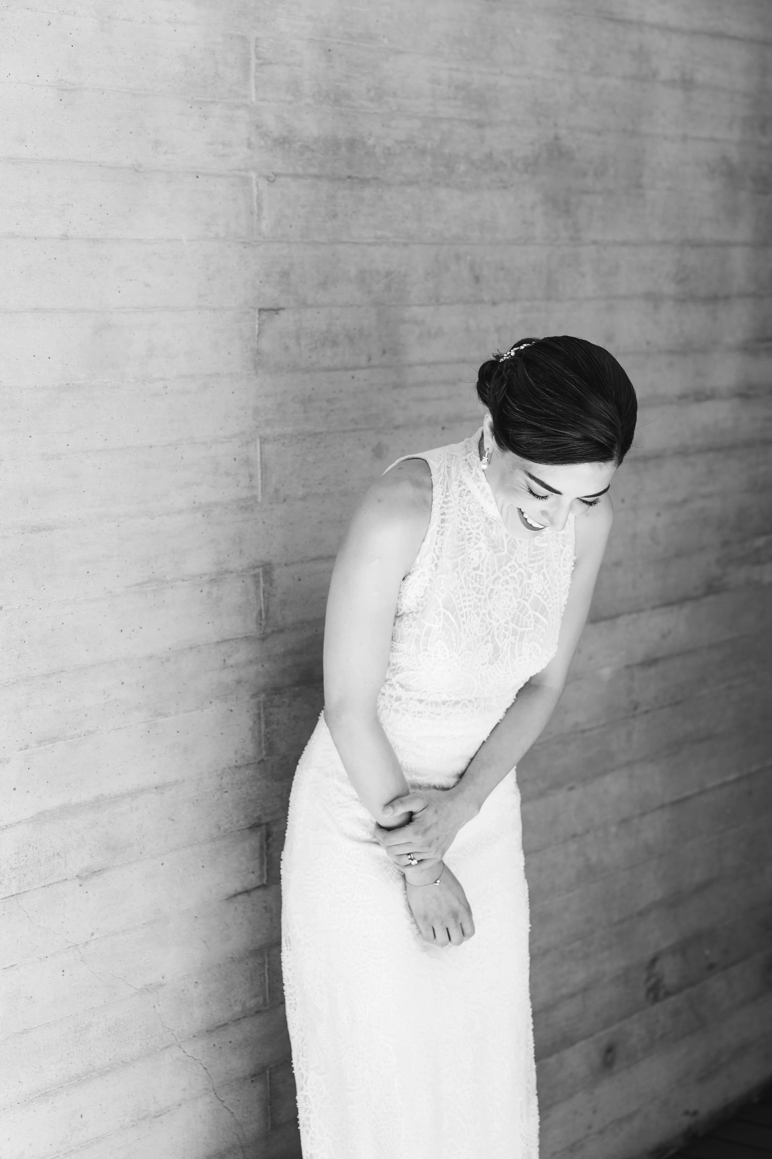 Black and white photo of a woman in a lace wedding dress, smiling and looking down, standing against a plain concrete wall.
