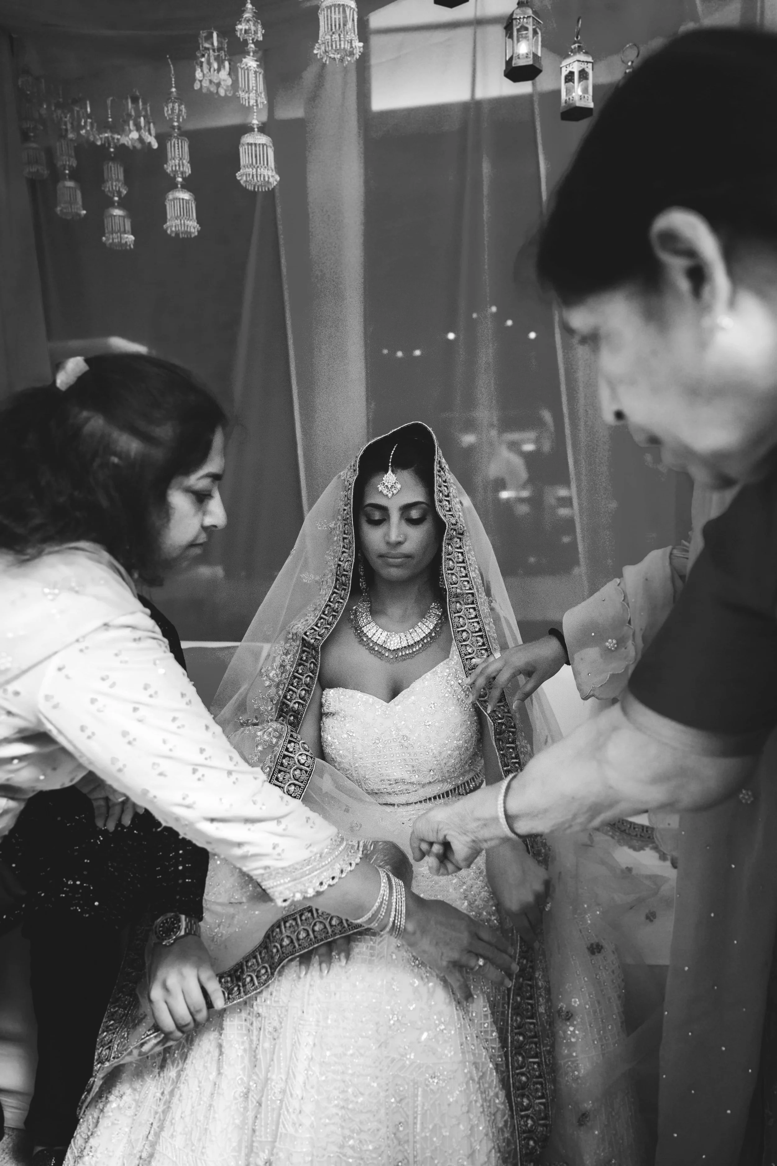 A bride in traditional Indian wedding attire, surrounded by four women and a man, possibly during a wedding ceremony. The women are dressed in white and black, and the man is in a dark suit. They are all holding the bride's hands, in a moment of ritu